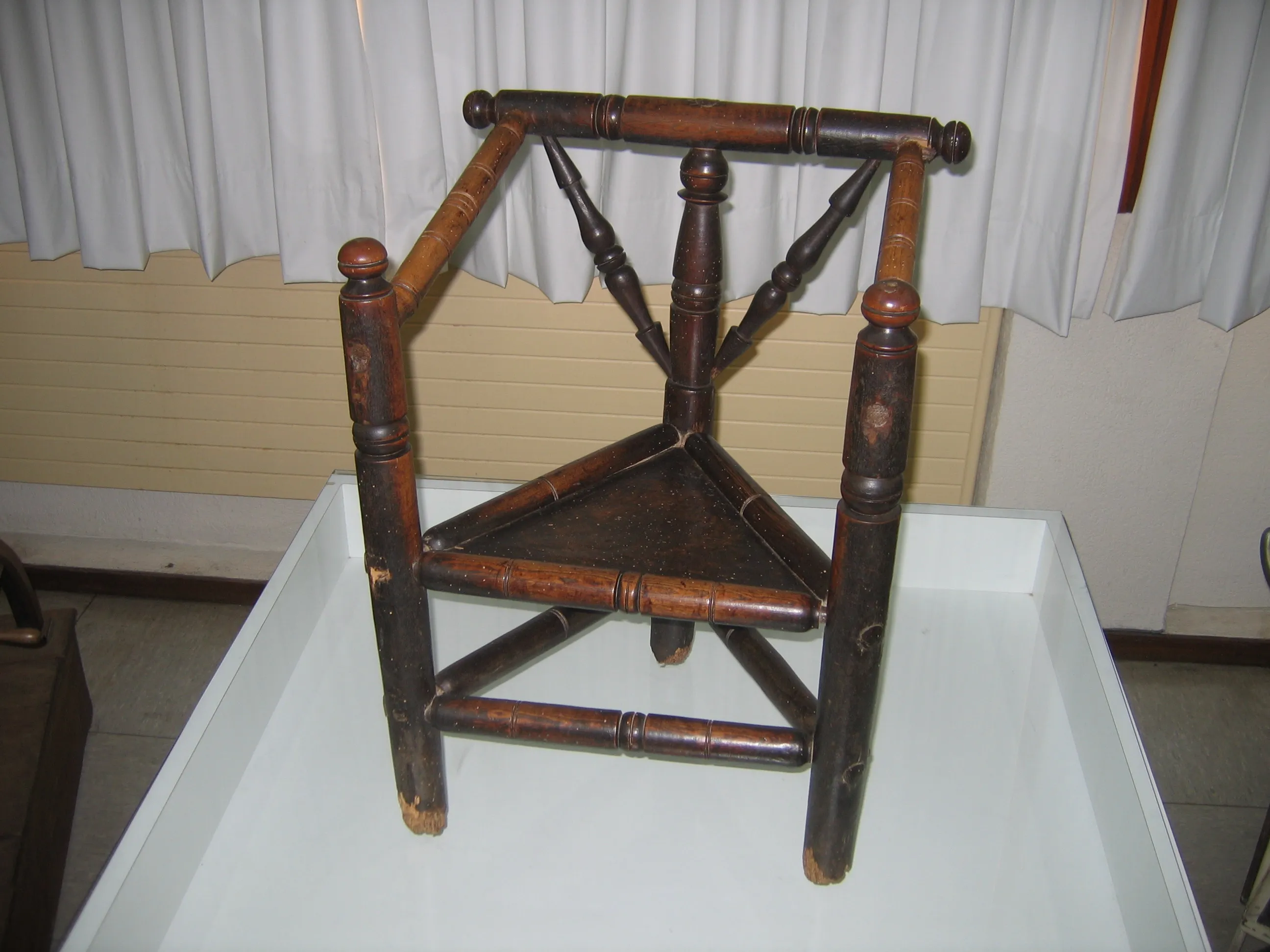 Wooden 18th-century labor chair with triangular seat and turned spindle backrest, displayed on a white pedestal against white curtains