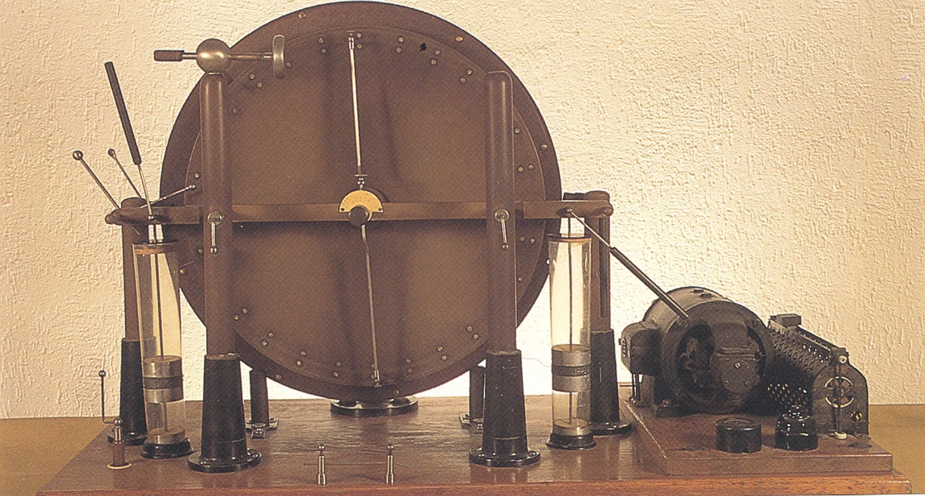 Wimshurst electrostatic generator circa 1900, a large circular disc apparatus with mechanical arms, glass cylinders, and a motor mounted on a wooden base against a plain wall
