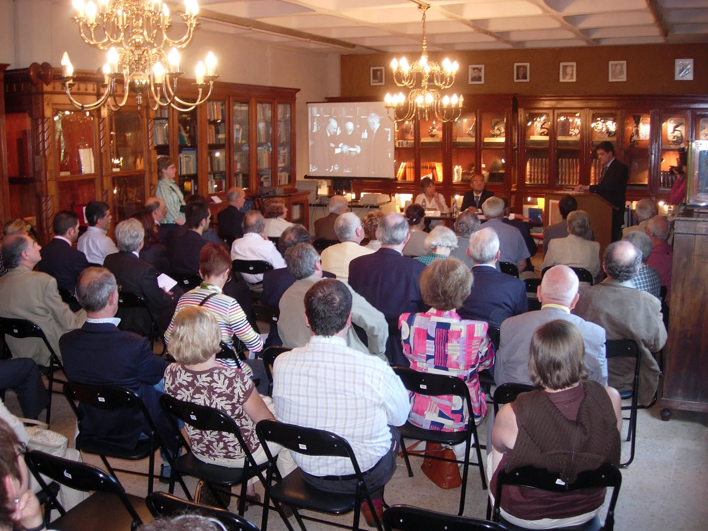 Audience seated in rows of chairs in a wood-paneled hall with ornate chandeliers and tall bookcases, watching a presentation on a projection screen
