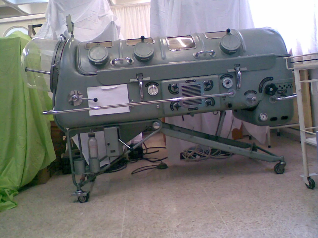 Large gray cylindrical steel lung iron lung medical device from the mid-20th century on a wheeled metal stand with dials, gauges, and control ports along its side