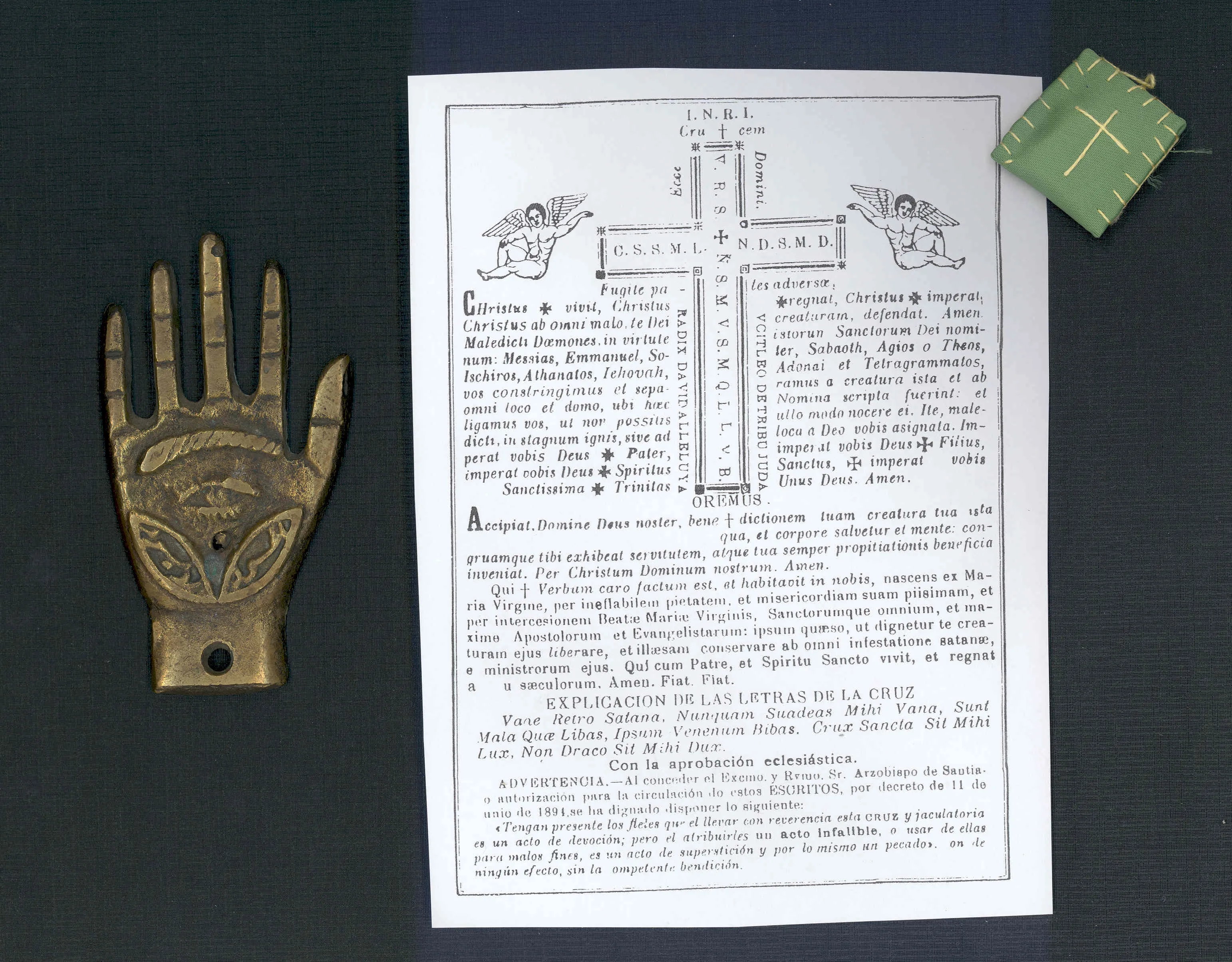 Bronzen amulet in de vorm van een hand met een oogmotief, tentoongesteld naast een gedrukt document met tekst en diagrammen en een klein groen tasje op een donkere ondergrond