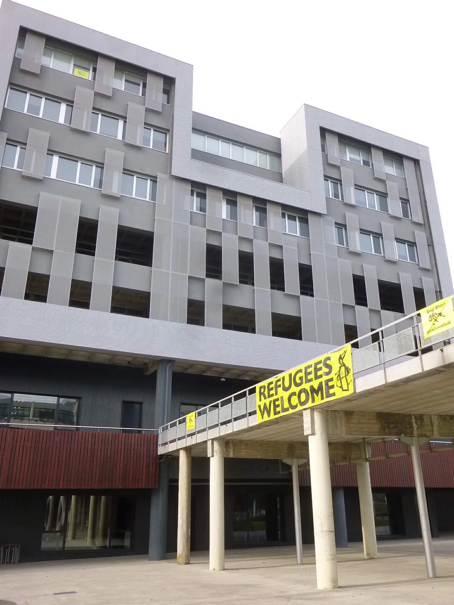 Gray multi-story concrete university building with a yellow 'Refugees Welcome' banner hanging from an elevated pedestrian walkway supported by columns