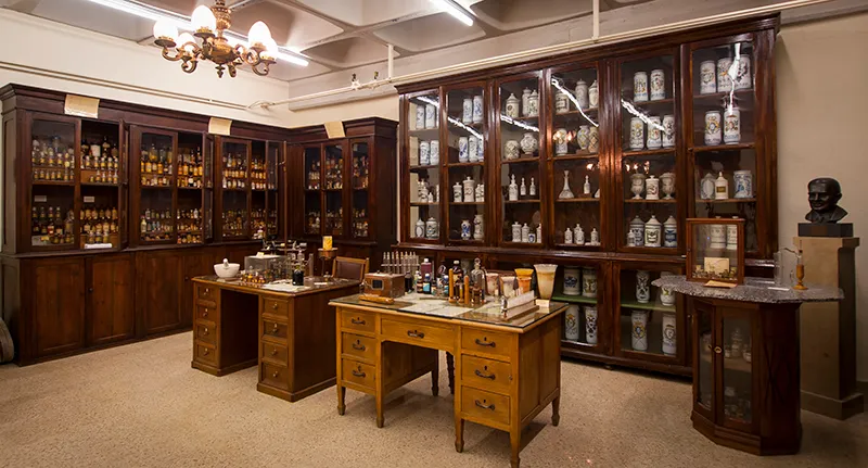 Interior of a historical pharmacy museum room with tall wooden display cabinets filled with glass bottles and jars, wooden work tables with medical instruments, and a chandelier overhead