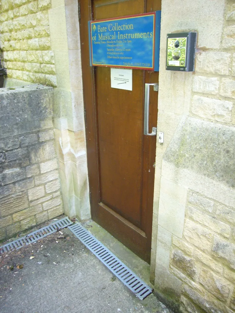 Wooden entrance door with a blue sign reading 'Bate Collection of Musical Instruments' set in a stone wall, with a metal drain grate on the ground.