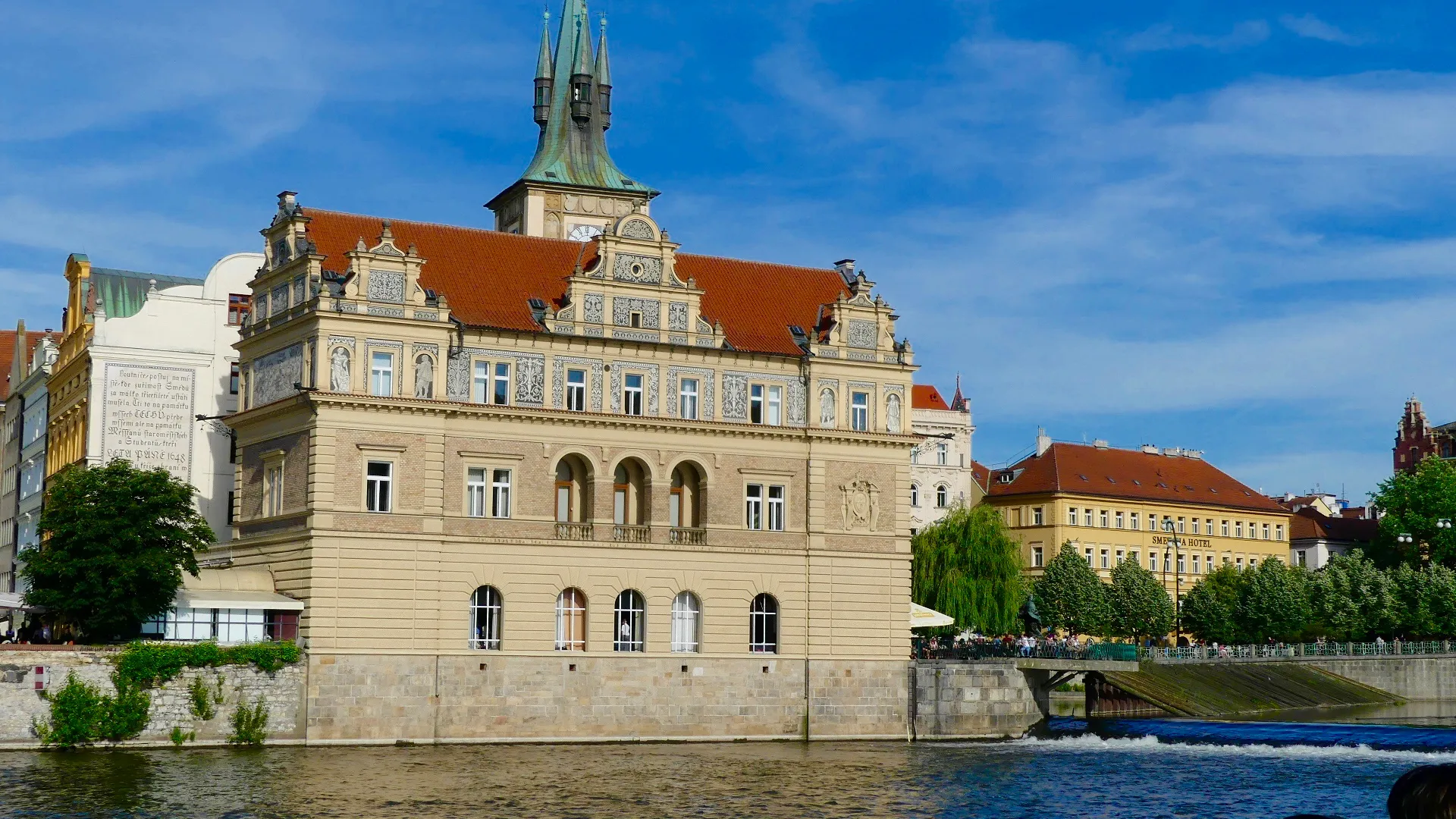 Budynek neorenesansowy z dachem z czerwonej dachówki i kamienną fasadą nad brzegiem rzeki pod błękitnym niebem