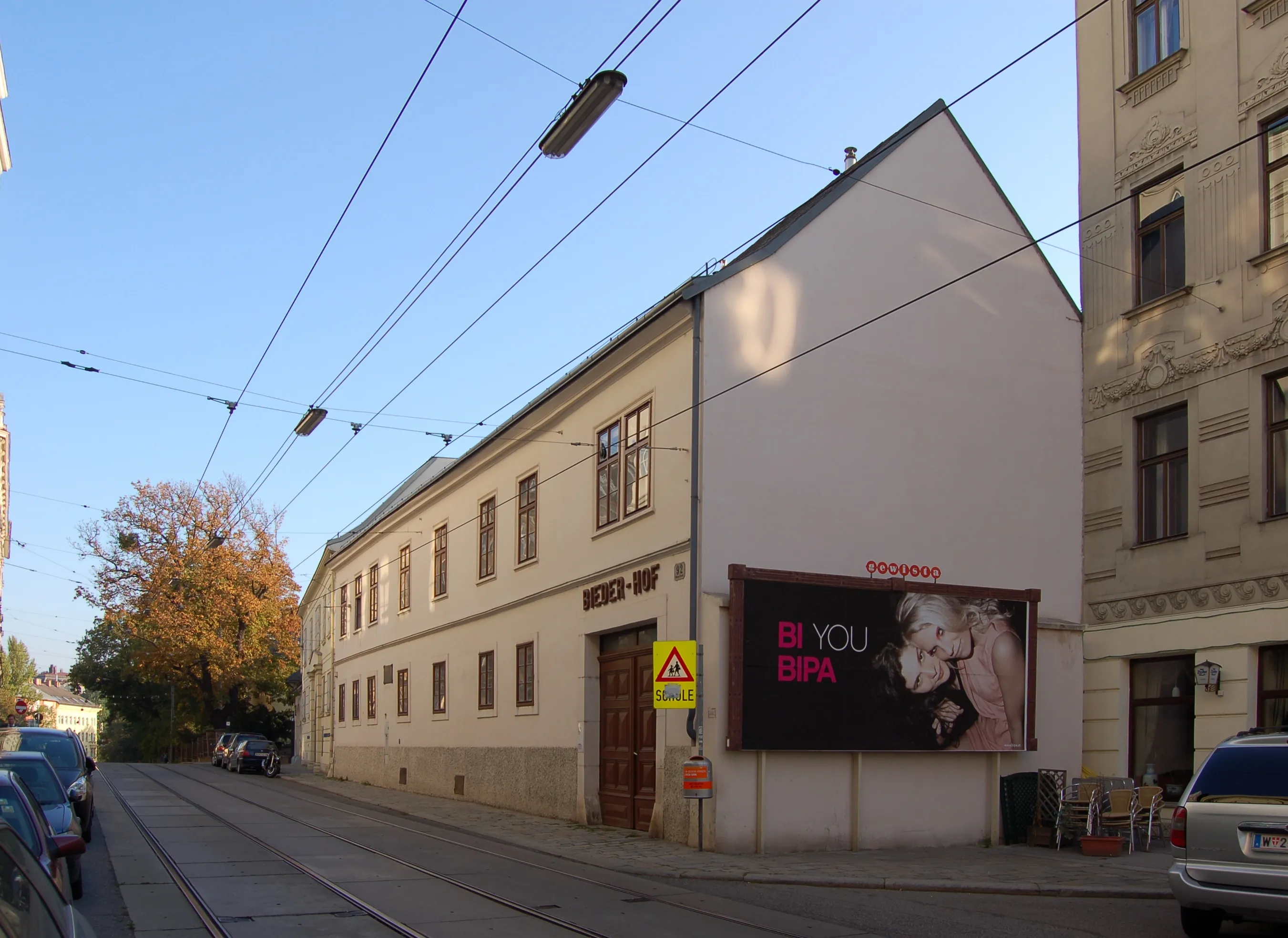 Biały budynek z dużą reklamą z boku, ulica z torami tramwajowymi i zaparkowanymi samochodami