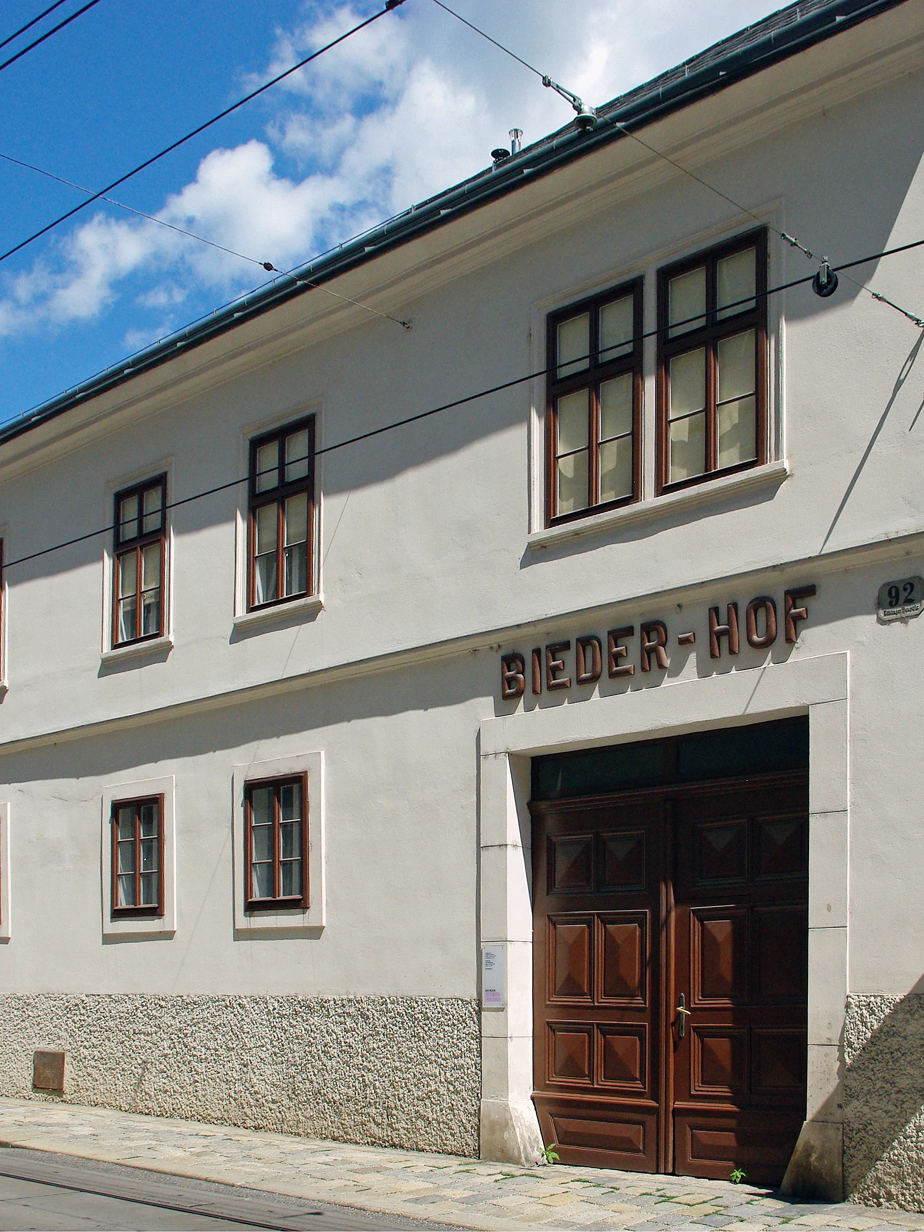 biały budynek z brązowymi drewnianymi drzwiami i znakiem BIEDER-HOF nad wejściem