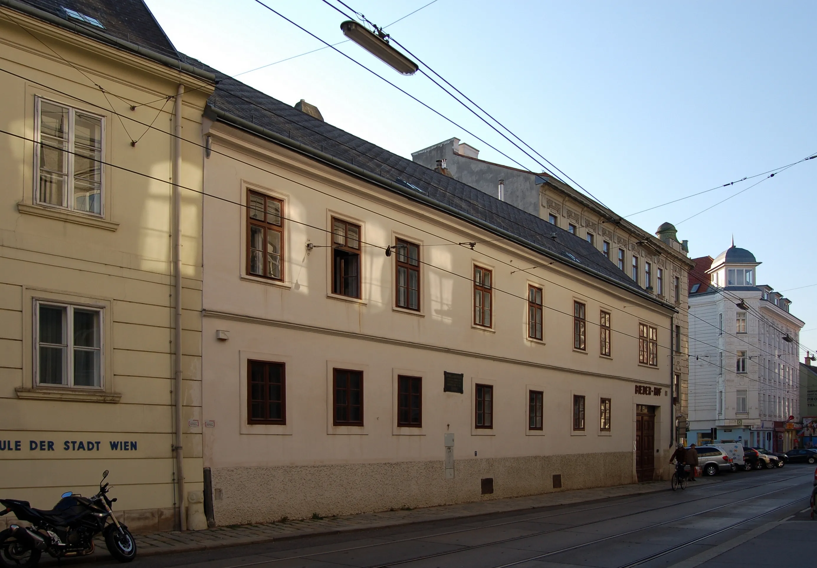 Bladożółty budynek z wieloma oknami i ciemnym dachem, położony przy ulicy z liniami tramwajowymi nad nią i zaparkowanym z przodu motocyklem.