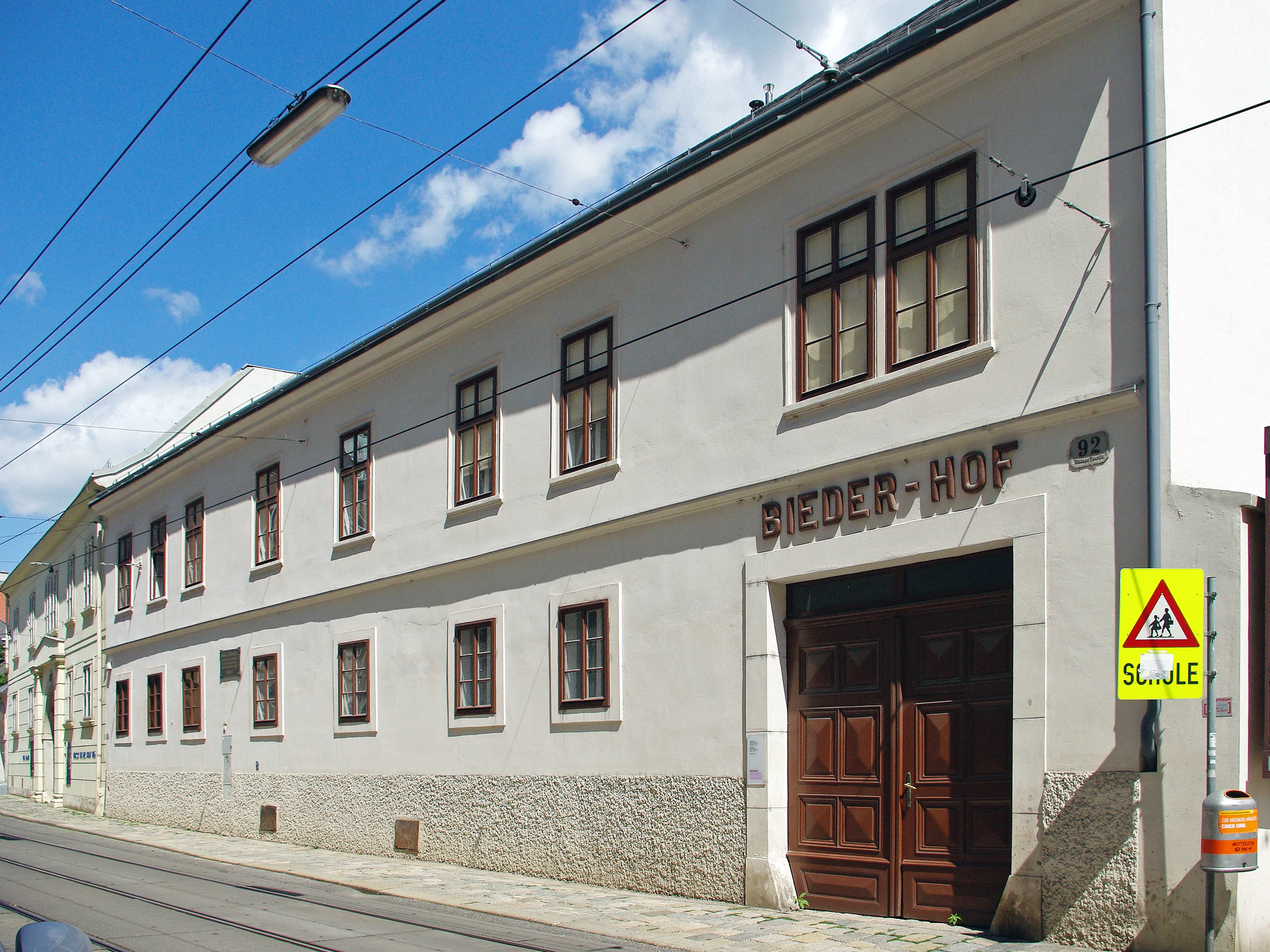 biały budynek z brązowymi oknami i drewnianymi drzwiami, napis BIEDER-HOF, błękitne niebo z chmurami