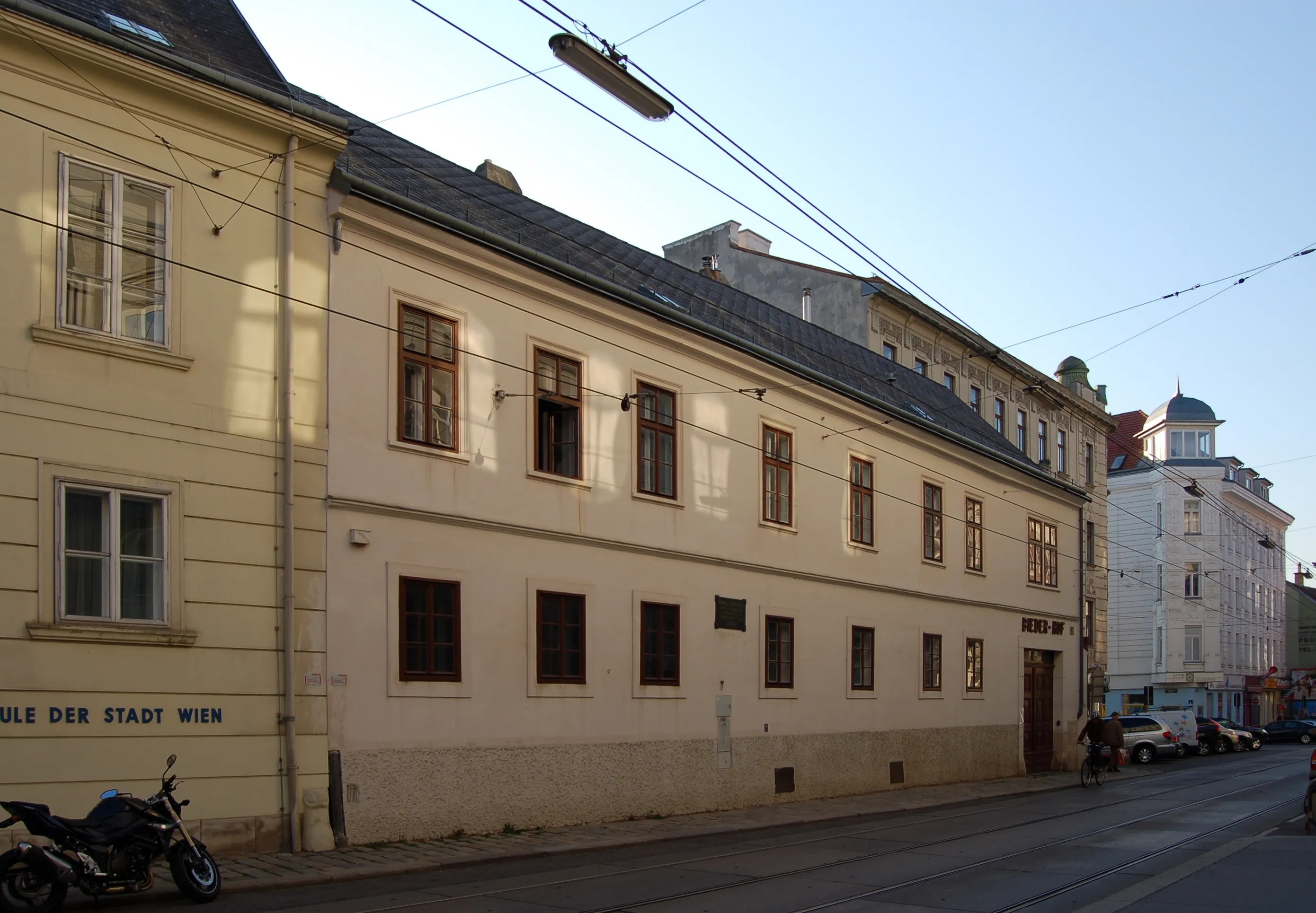 Jasny budynek z wieloma oknami i znakiem na scianie, polozony na rogu ulicy w Wiedniu.