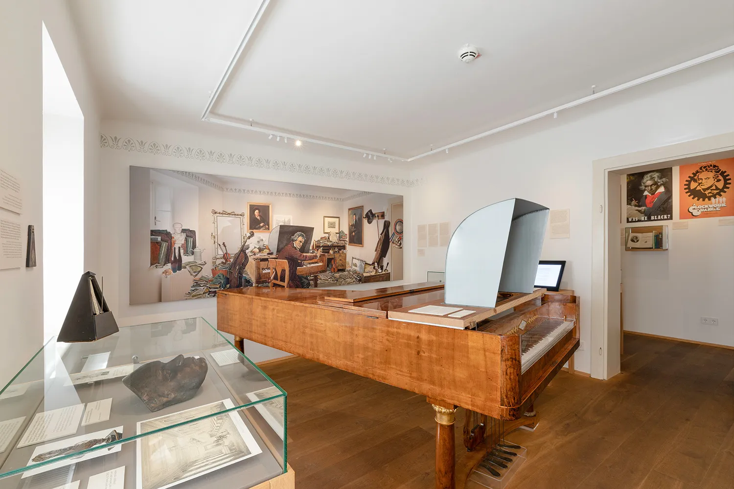 Un fortepiano expuesto en una galería de museo con un gran mural en la pared y vitrinas que contienen artefactos.