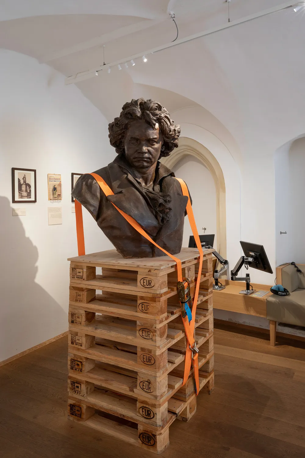 Busto de bronce de Beethoven sobre palets de madera con correas naranjas en una galería de museo