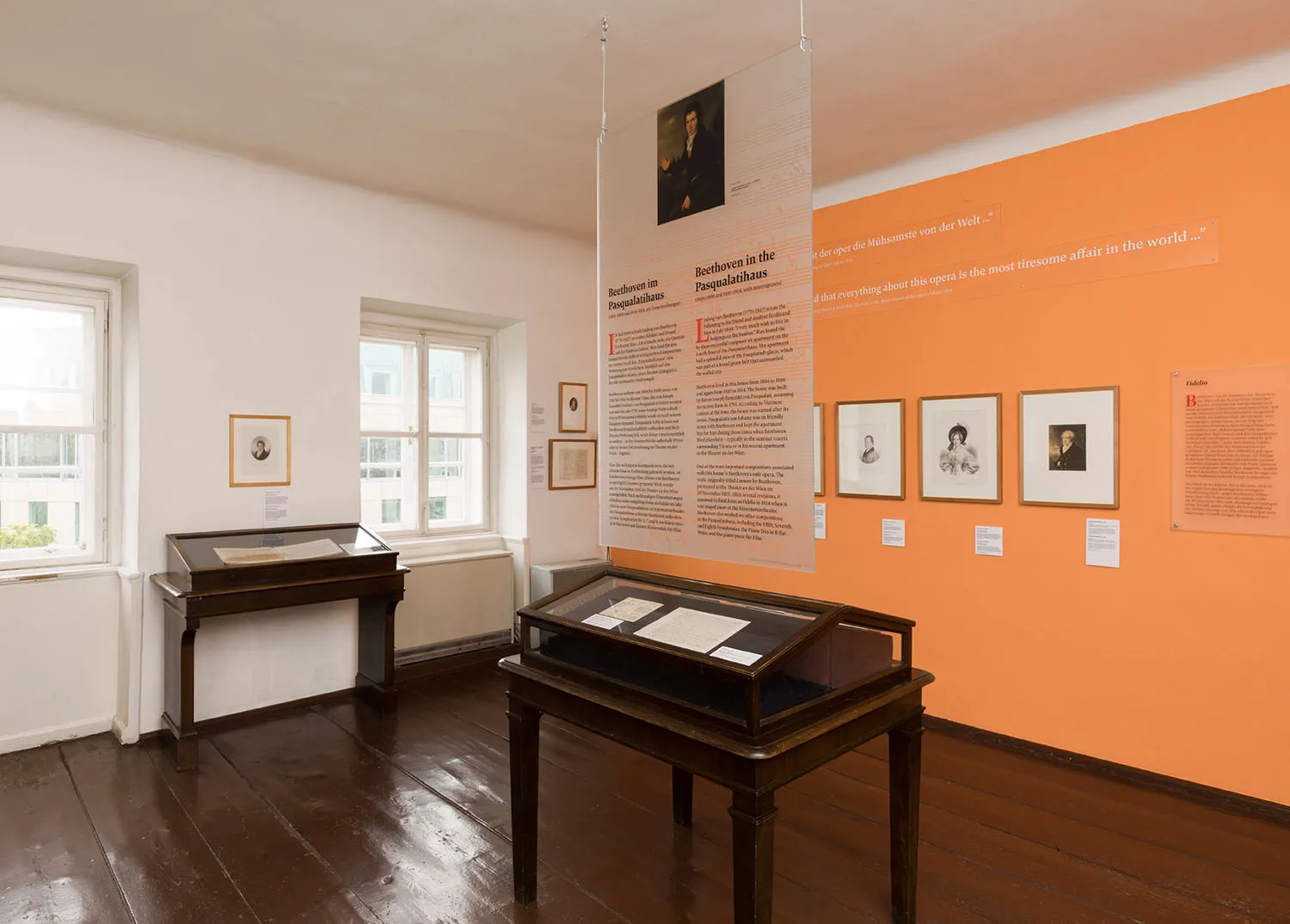 Interior of Beethoven Pasqualatihaus with wooden display cases, framed documents, and an orange wall with informational text.