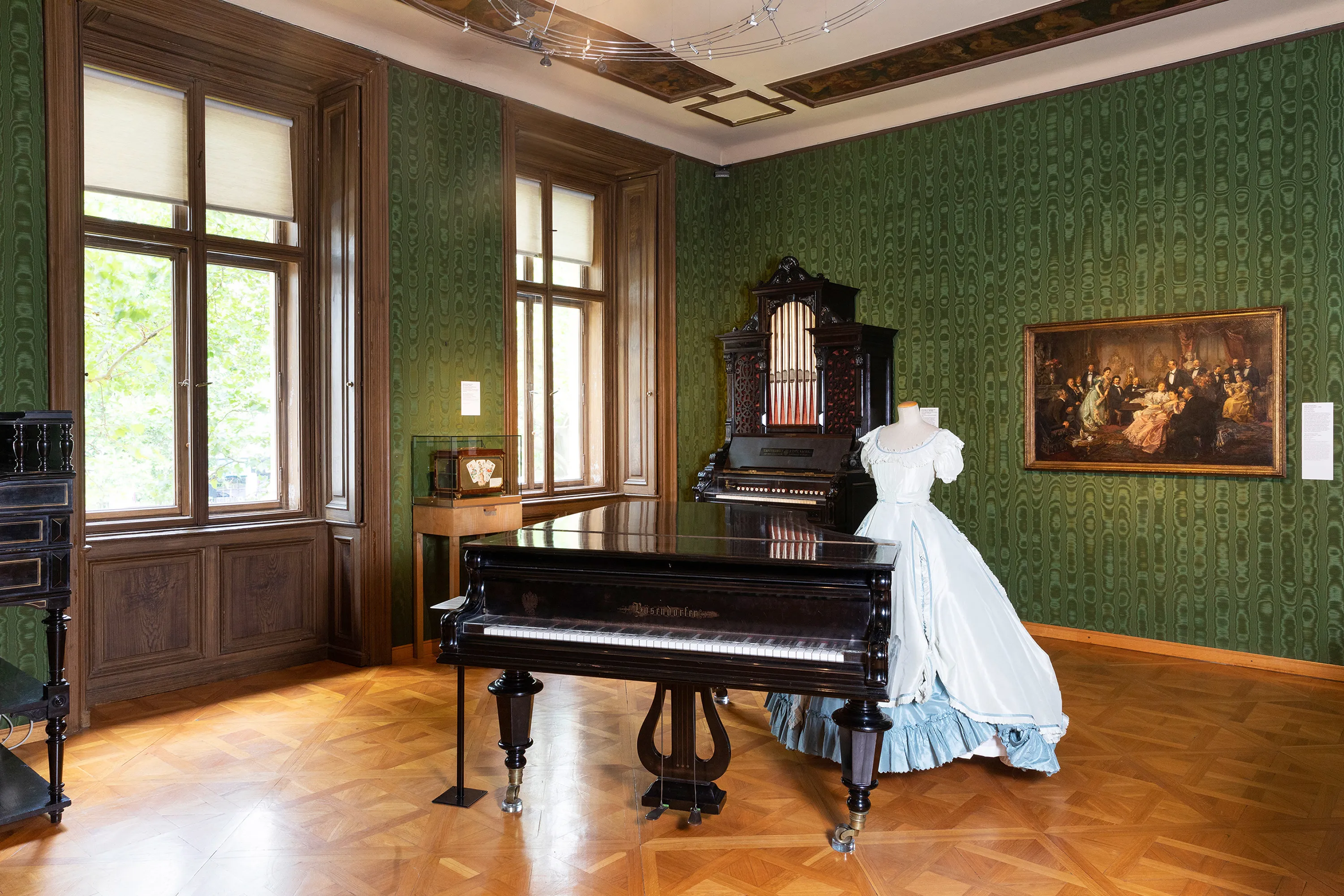 A grand piano in a room with green wallpaper, wooden floors, and a mannequin in a white dress.