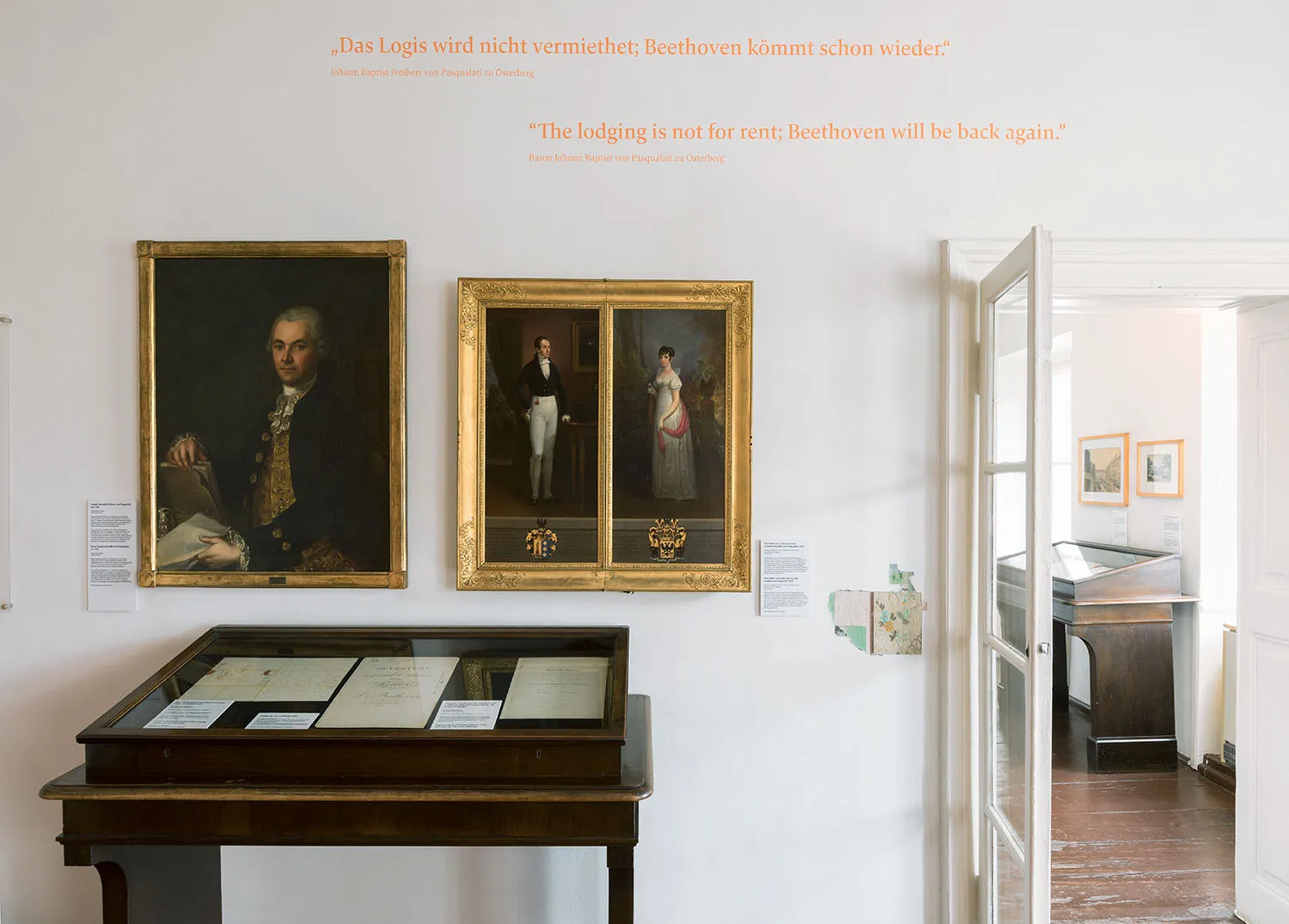 Interior of Beethoven Pasqualatihaus with two framed portraits on a white wall and a display case in the foreground