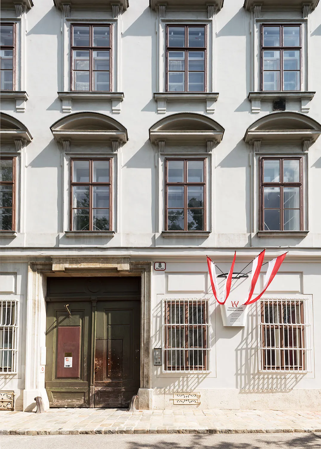A light-colored building facade with arched windows, a green entrance door, and a red and white banner with a 'W' logo
