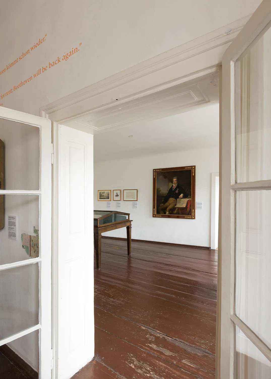Pasillo interior con suelos de madera y paredes blancas, que conduce a una galería con un retrato de Beethoven y una vitrina.