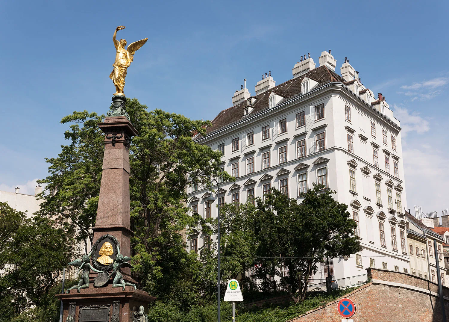 Un edificio clásico blanco con muchas ventanas y un tejado oscuro, con una estatua dorada sobre un pedestal de piedra en primer plano.