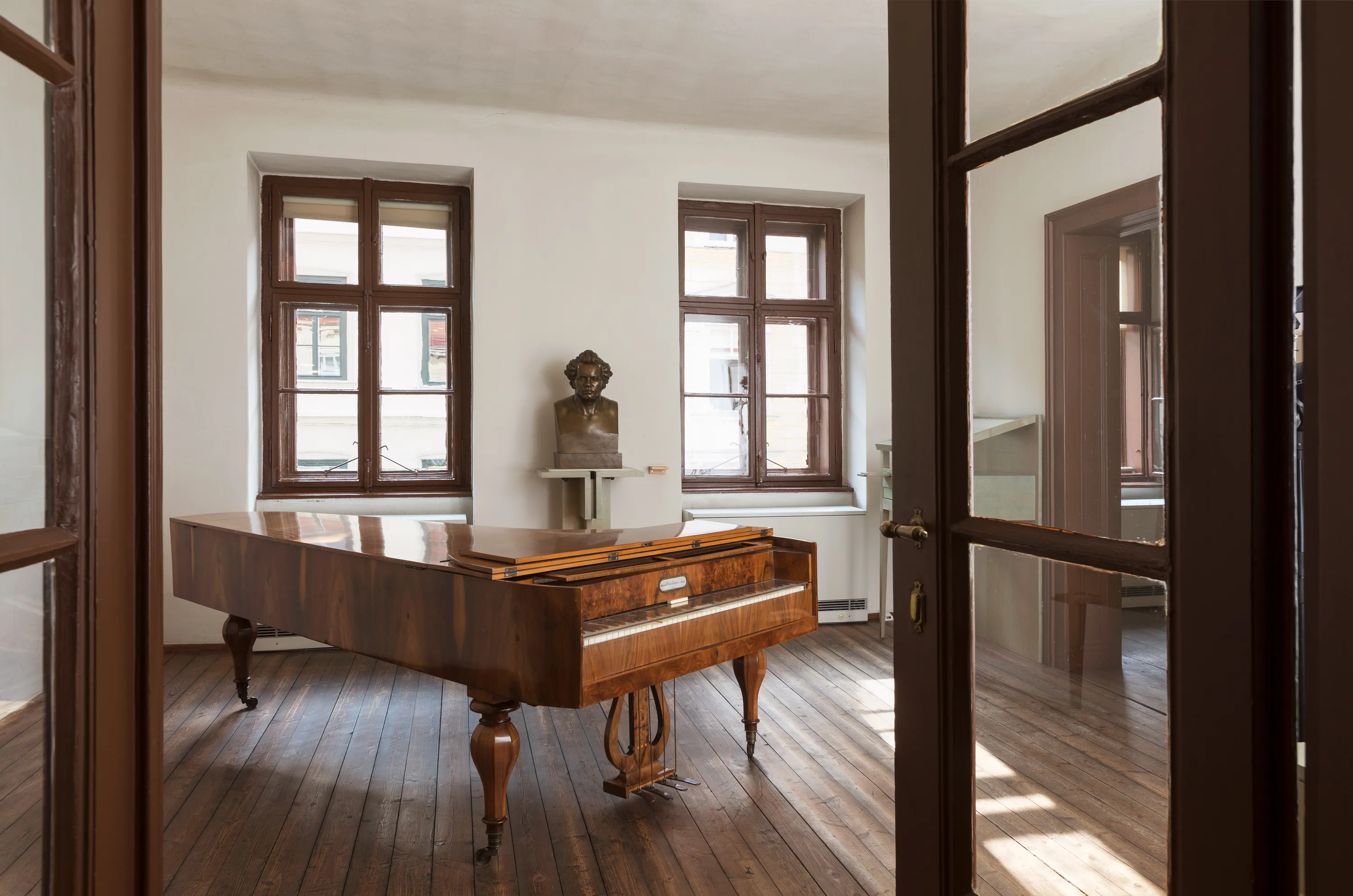 A wooden grand piano in a room with wooden floors and two windows, viewed through a doorway with glass panels.