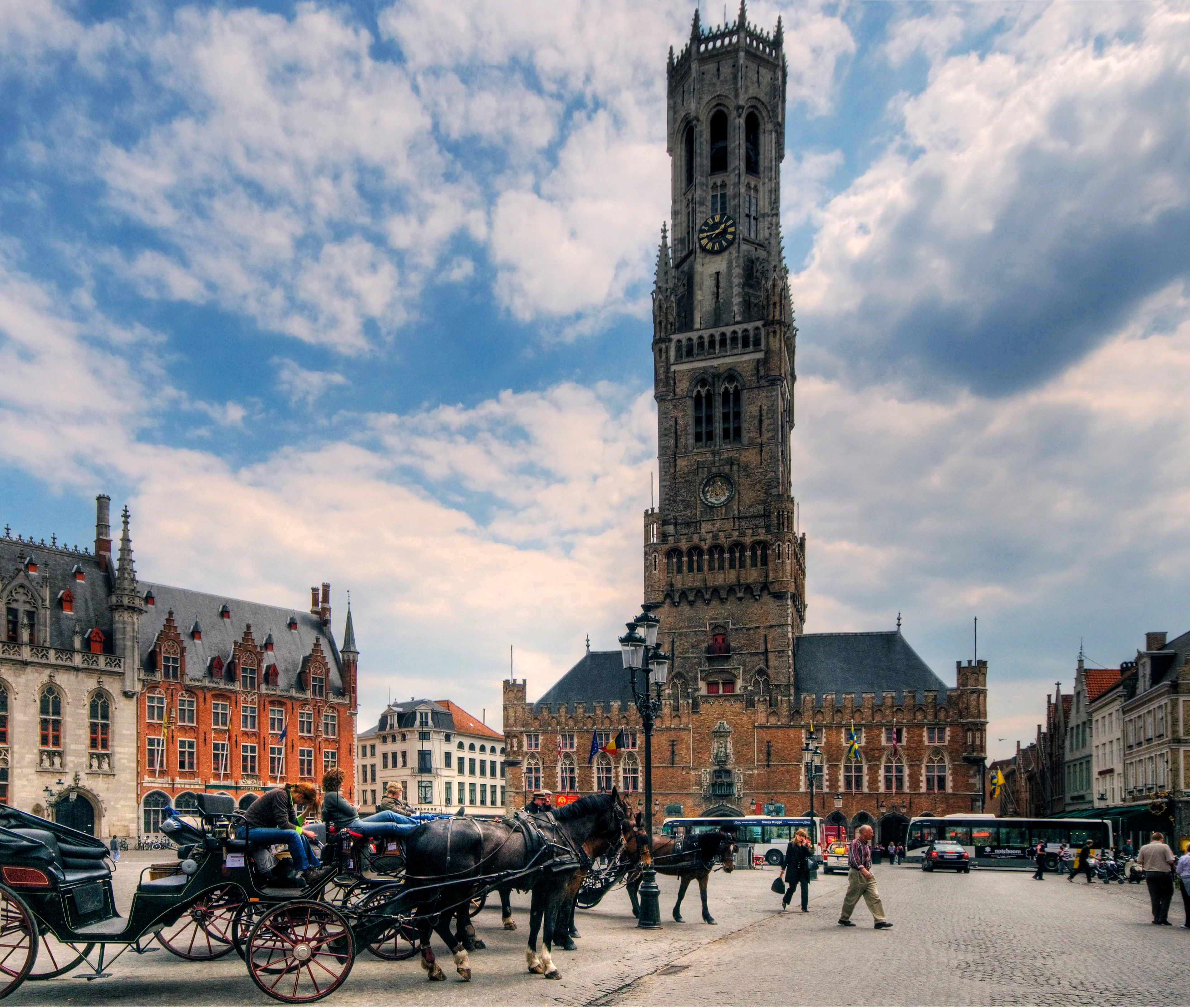 The Belfry of Bruges tower rises above Market Square with horse-drawn carriages and pedestrians in the foreground under a cloudy blue sky