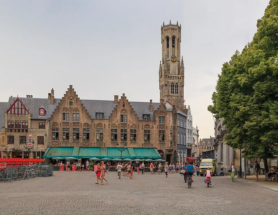 Szeroki widok na Rynek w Brugii, ukazujący wieżę Belfort oraz budynek Sądu Prowincjonalnego z zielonymi markizami, z ludźmi spacerującymi i jeżdżącymi na rowerach po bruku.