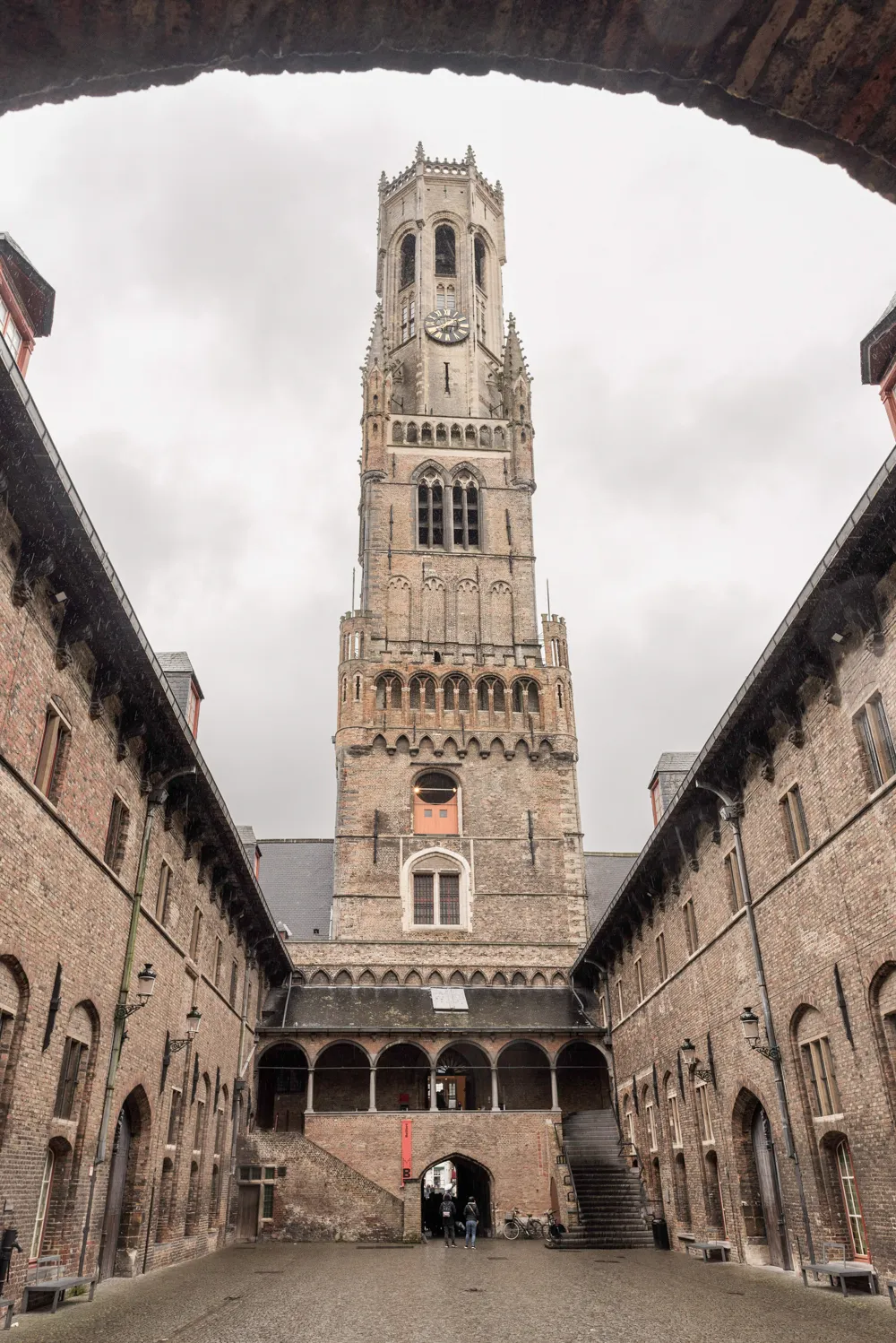 The medieval Belfort tower of Bruges rising above brick courtyard buildings, viewed through a stone archway under an overcast sky.