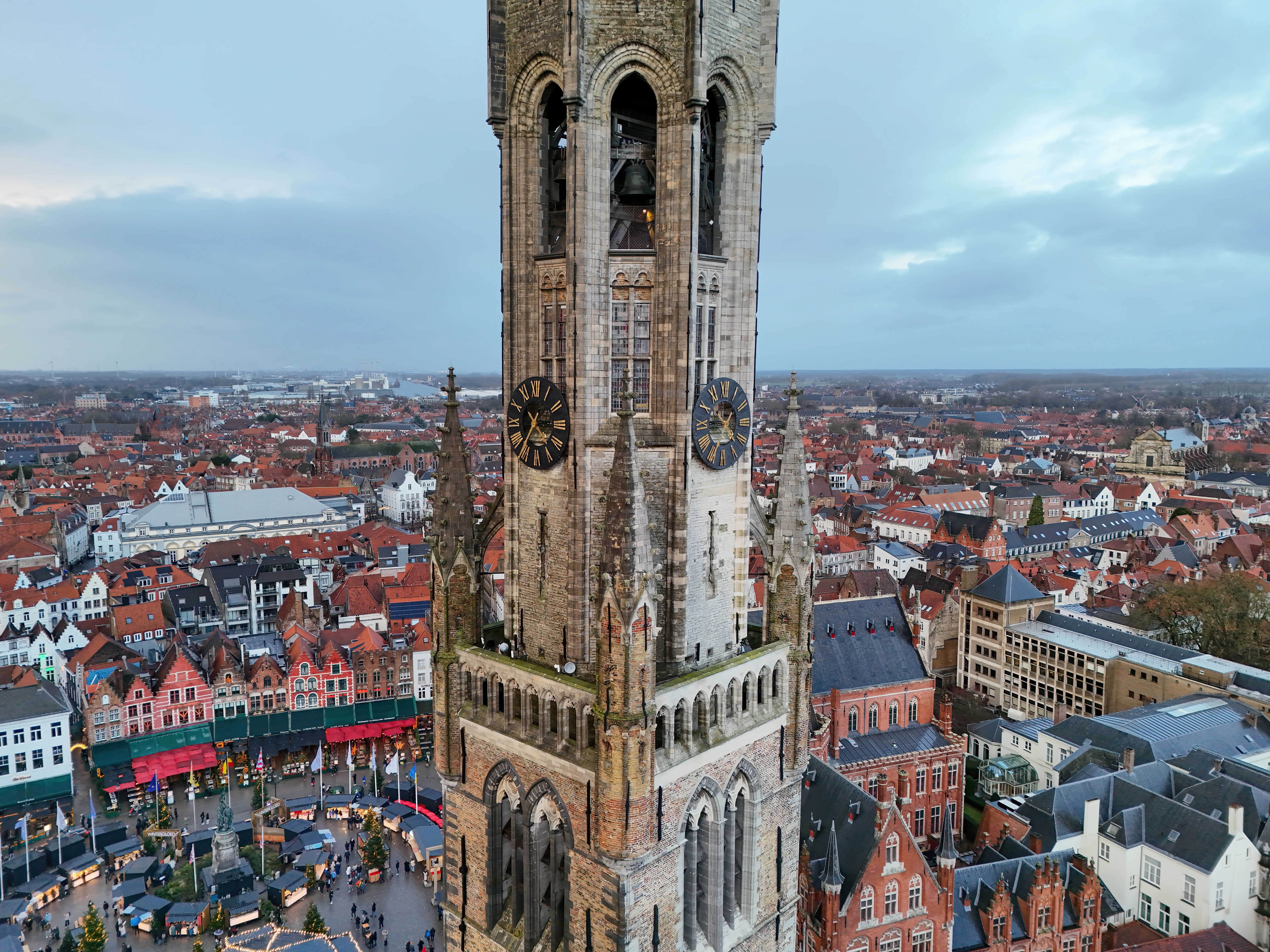 Aerial view of the Belfry of Bruges tower with clock faces on its stone facade, rising above the red-roofed cityscape of Bruges