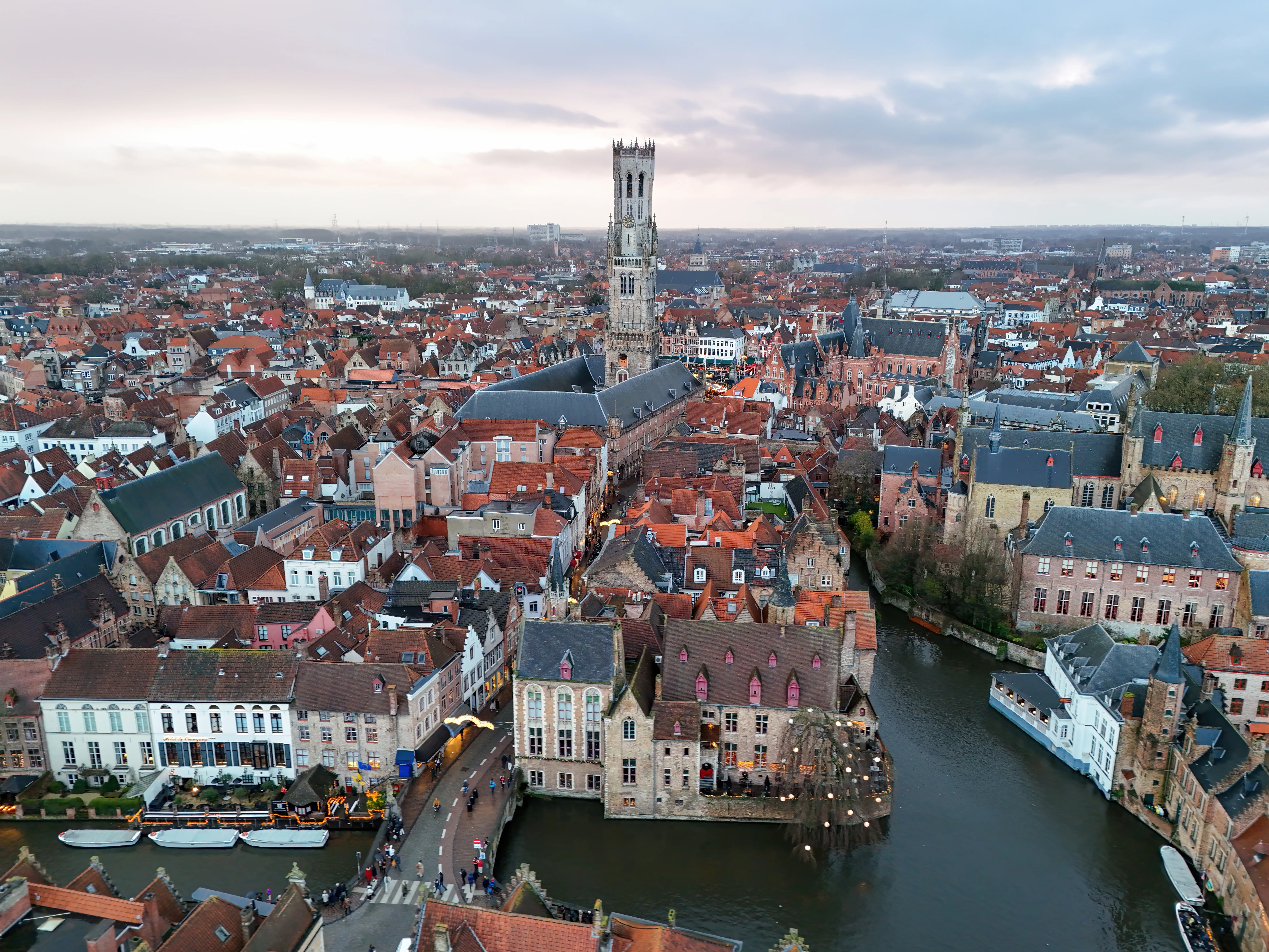 Panoramiczny widok z lotu ptaka na krajobraz Brugii o zmierzchu, ze średniowieczną wieżą Belfort wznoszącą się ponad dachami z czerwoną dachówką i kanałem wijącym się przez zabytkowe centrum.