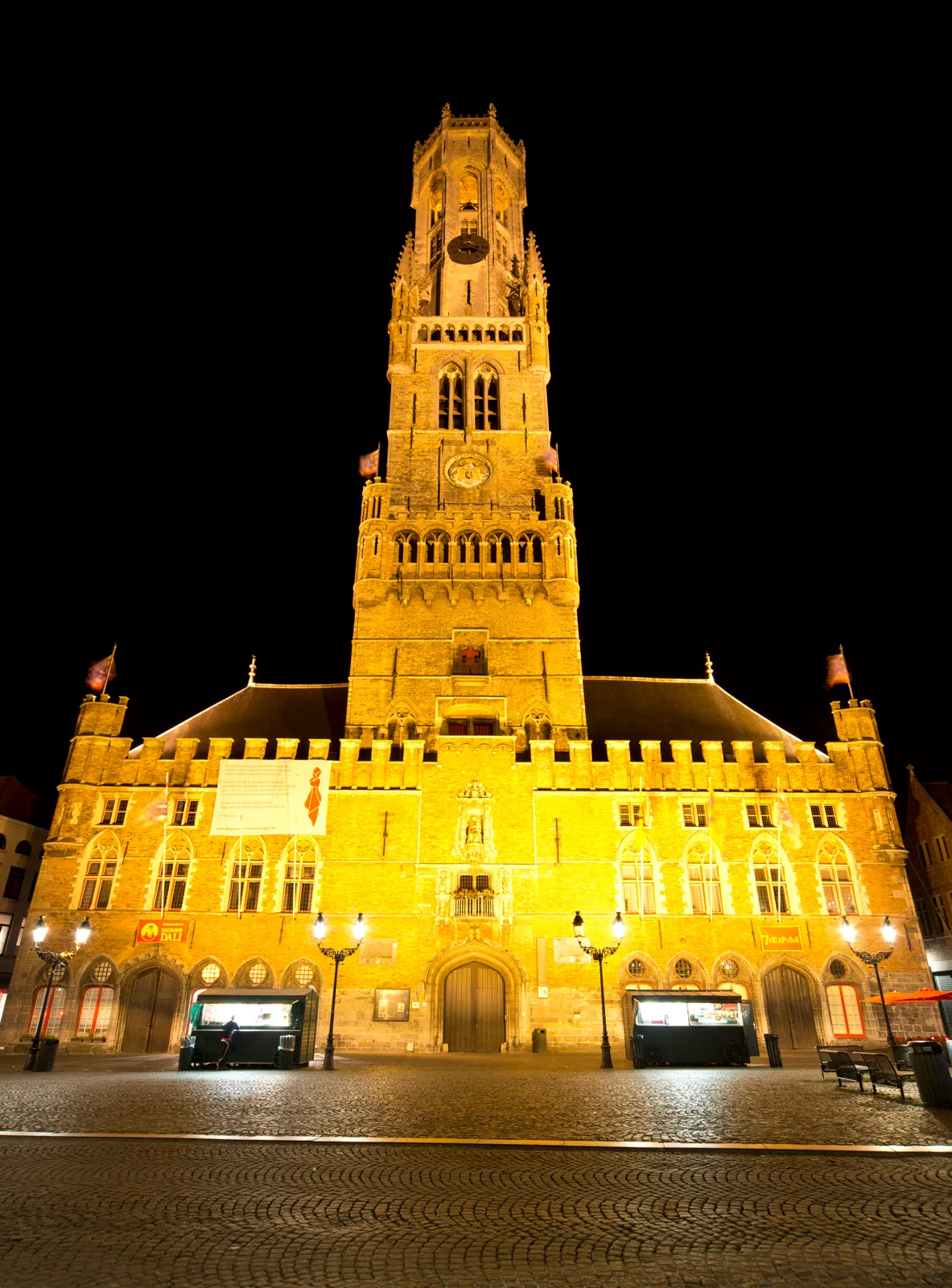 Illuminated medieval belfry tower rising above a brick building facade at night with cobblestone square and street lamps in the foreground