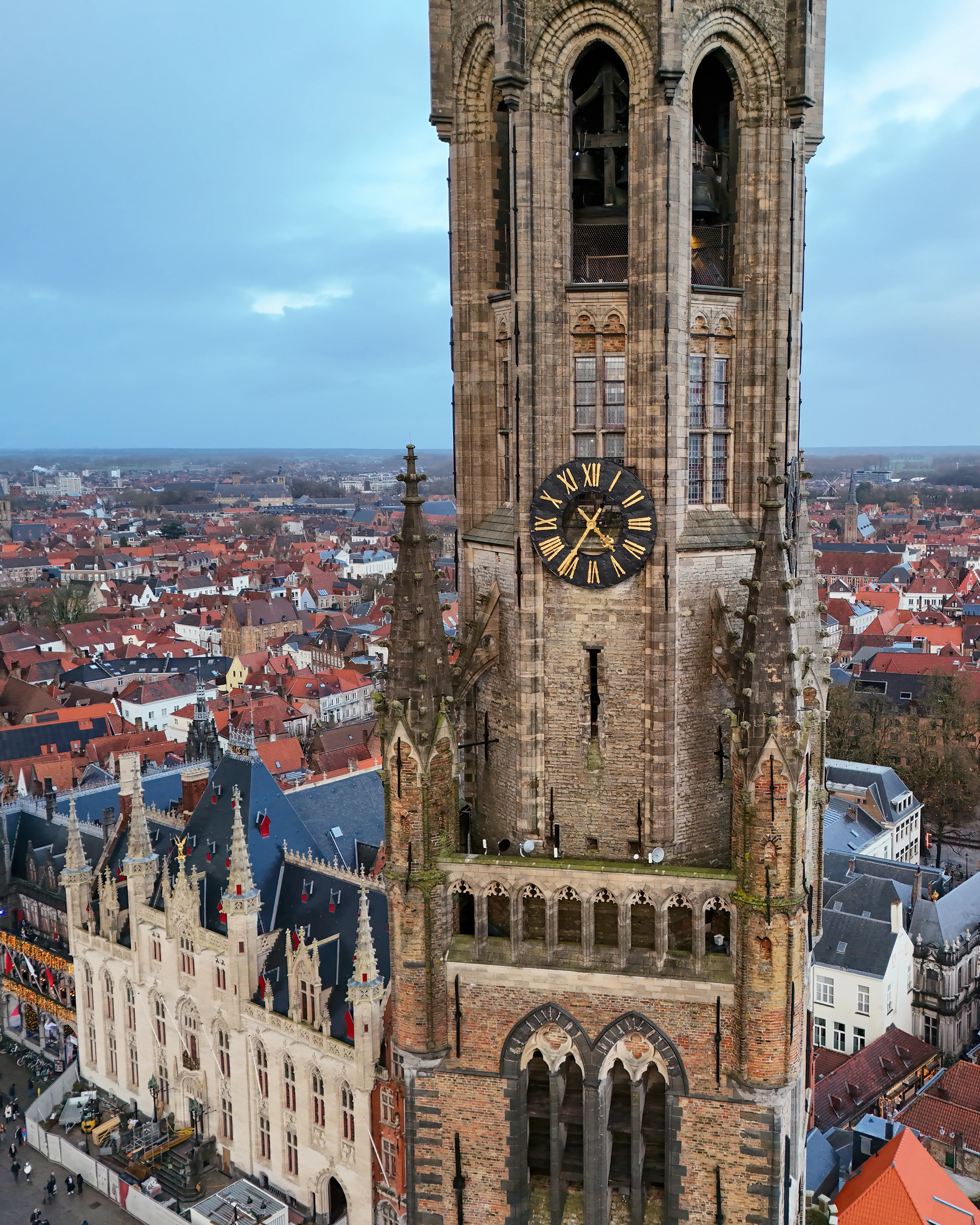 Aerial view of the Belfry of Bruges showing the tall brick clock tower with a Roman numeral clock face, Gothic spires, and the red-roofed cityscape of Bruges stretching into the distance