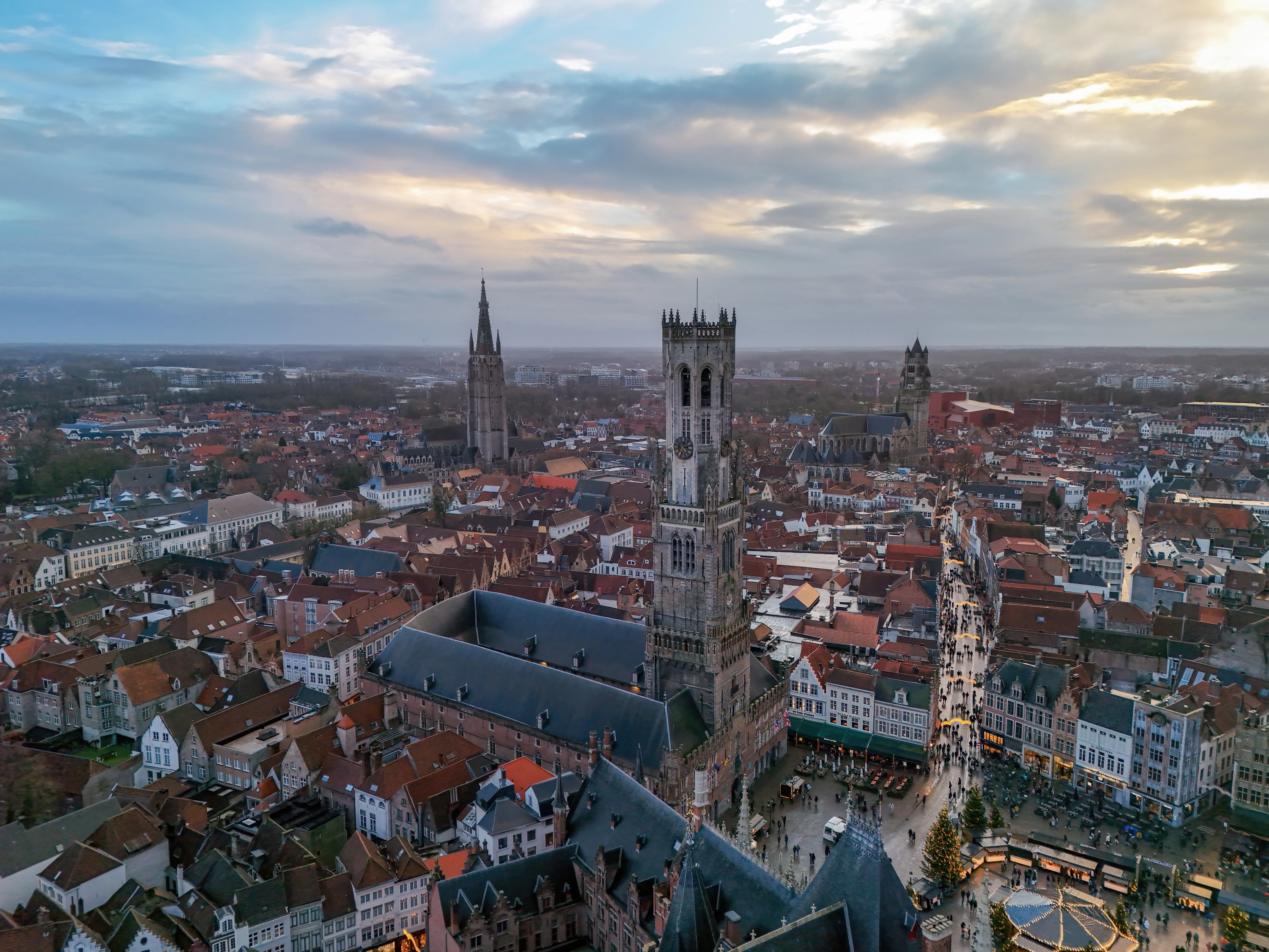 Aerial view of the Belfry of Bruges tower rising above the red-roofed historic city center and Market Square under a cloudy sky.