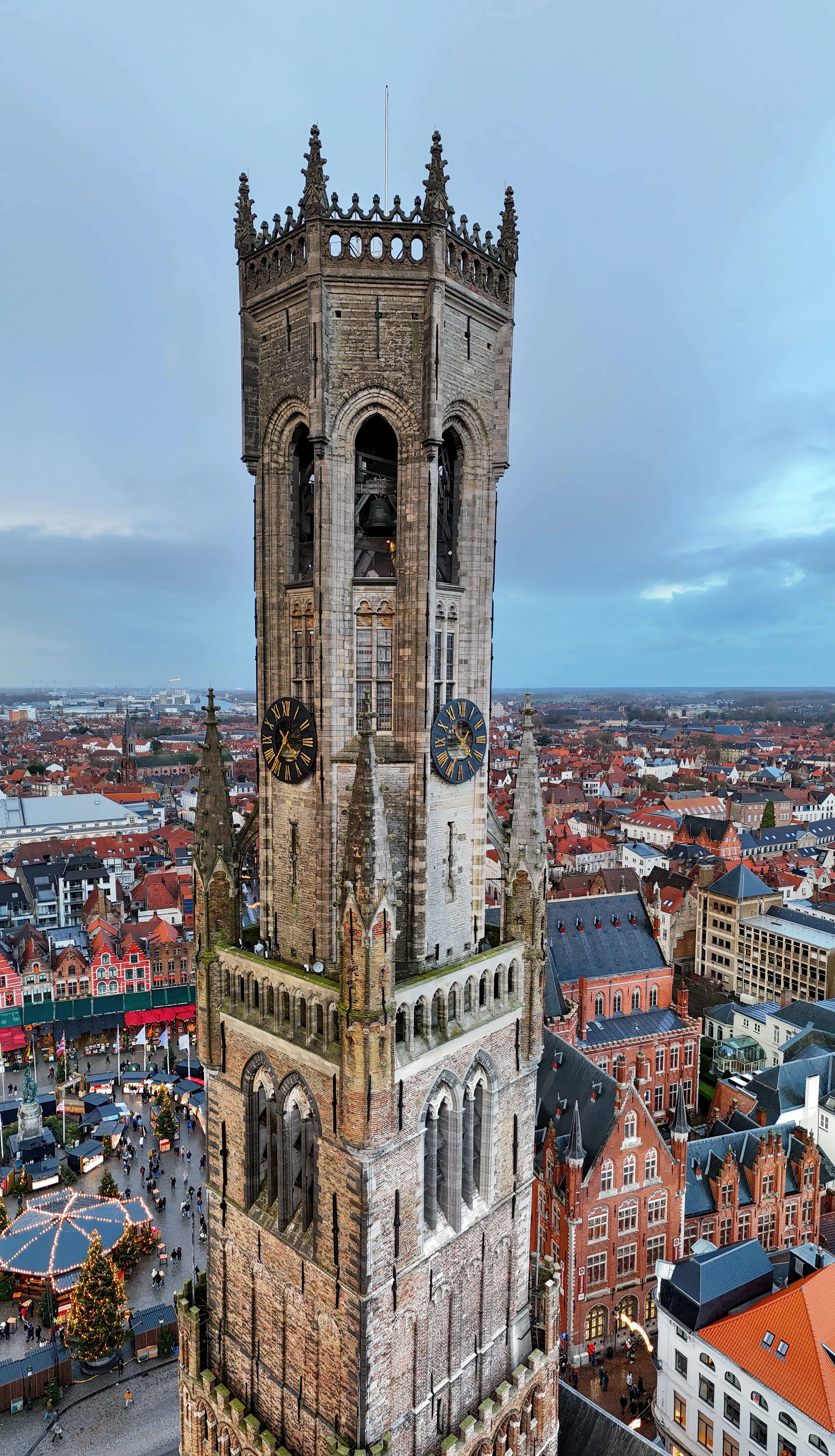 Aerial view of the Belfry of Bruges tower showing clock faces, bell openings with visible bells, and Gothic spires rising above the red-roofed cityscape and Market Square below