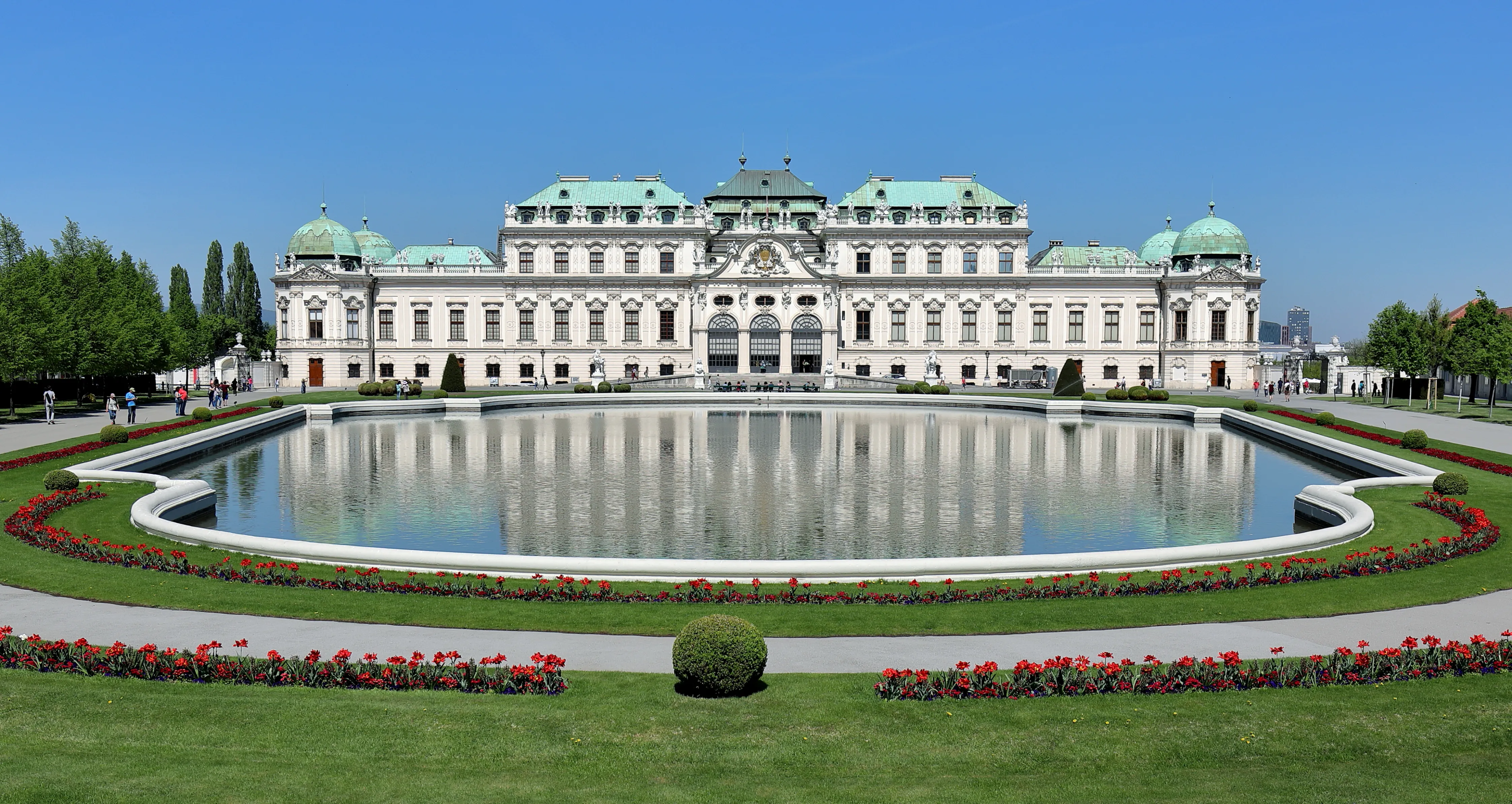 Pałac Belvedere z dużym basenem lustrzanym, zielonymi trawnikami i czerwonymi kwiatami przed białym barokowym budynkiem z zielonymi kopułami pod błękitnym niebem