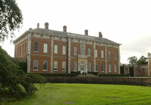 Red brick country house with white-framed windows, multiple chimneys, and a central entrance, viewed from a green lawn with trees to the left.