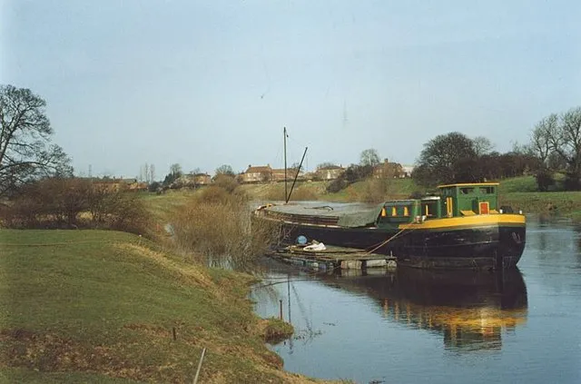 Green and yellow barge moored on the River Ouse with grassy banks and village houses in the background near Beningbrough