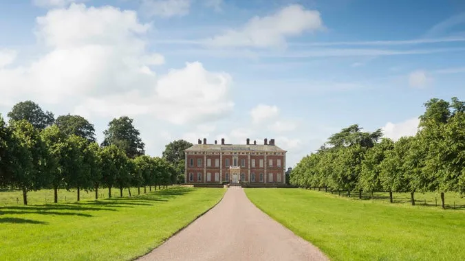Red-brick country house viewed down a long tree-lined path on a sunny day with blue sky