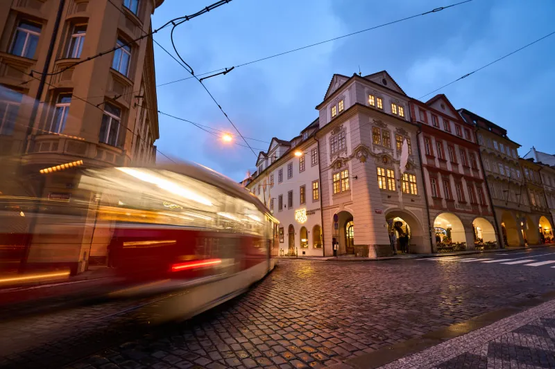 Budynek z XVIII wieku z oświetlonymi oknami i łukowym parterem, widziany z ulicy z poruszającym się tramwajem na pierwszym planie.