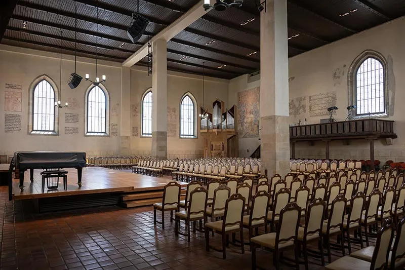 Interior de la Capilla de Belén con filas de sillas, un escenario con un piano, ventanas arqueadas y un techo de madera