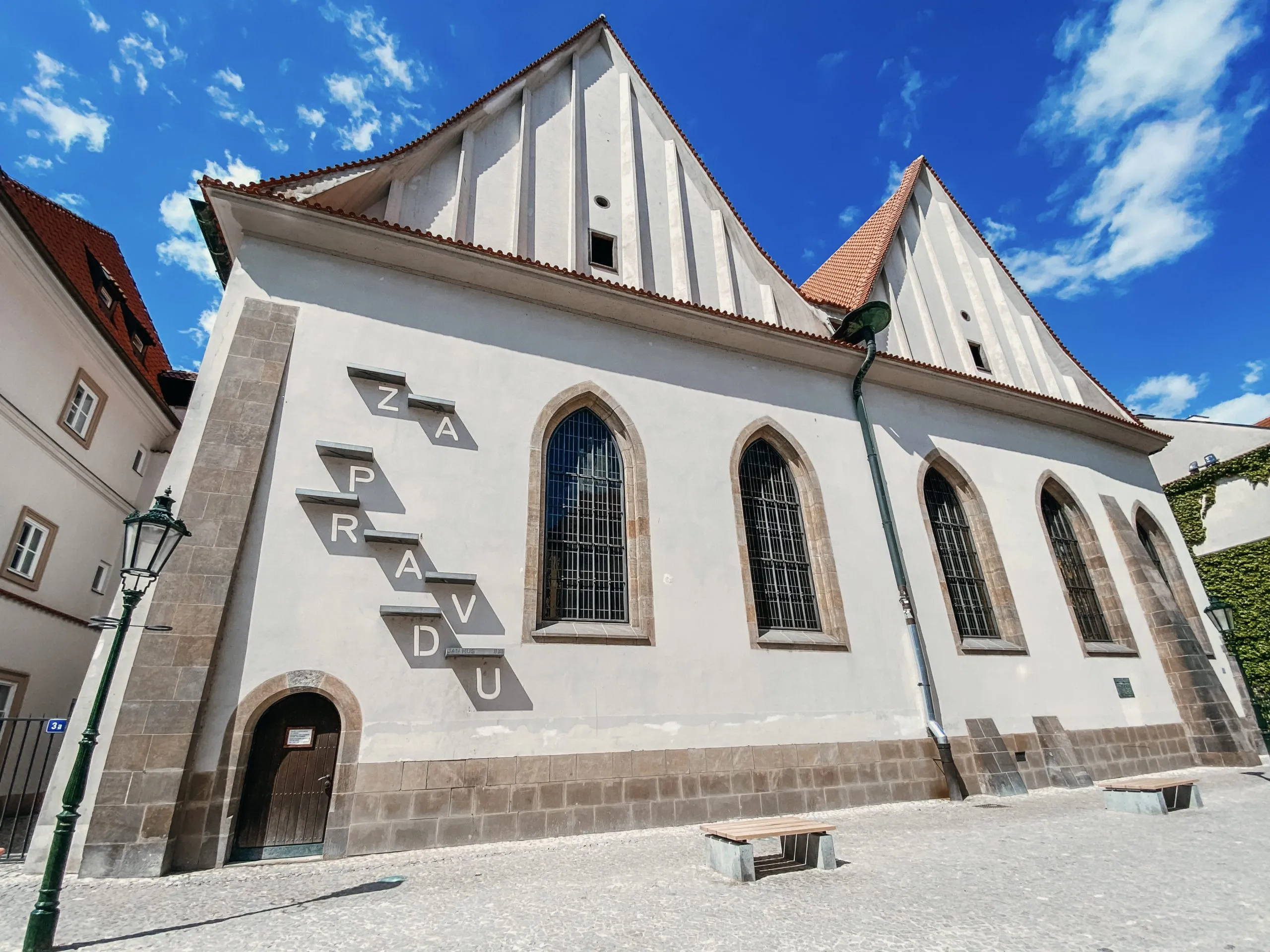 Una capilla blanca de estilo gótico con ventanas arqueadas y fachada de piedra, que presenta la palabra 'ZAPRAVDU' en letras grandes, bajo un cielo azul con nubes.
