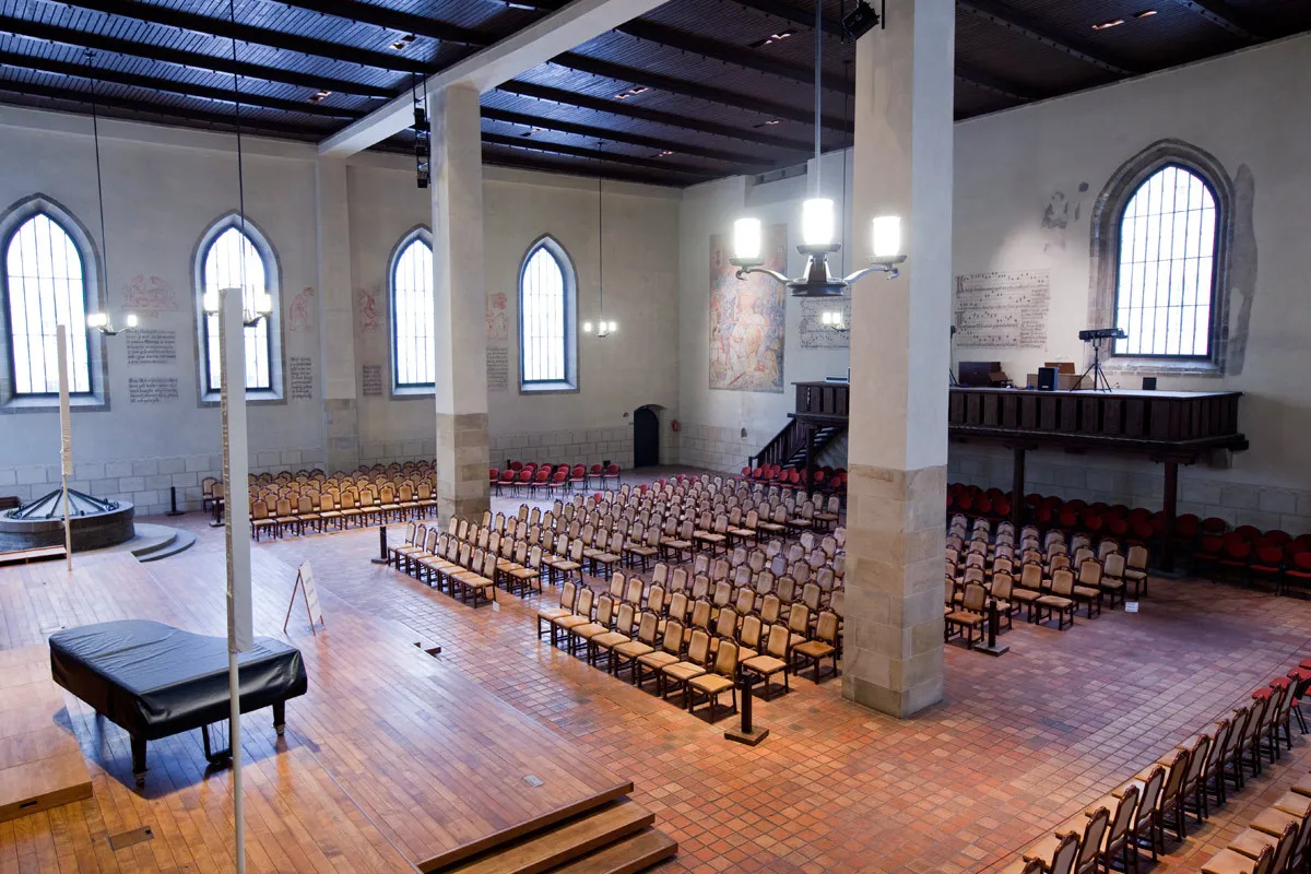Interior de la Capilla de Belén con hileras de sillas de madera, ventanas arqueadas y vigas de madera expuestas en el techo