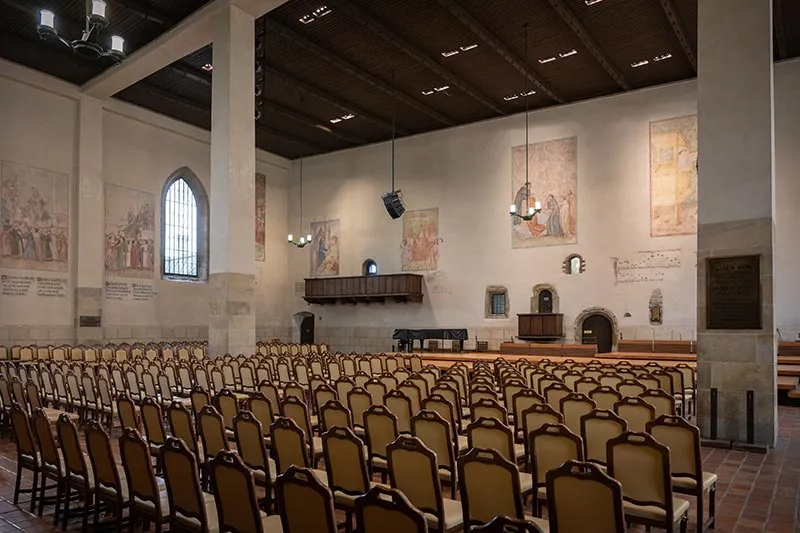 Interior de la Capilla de Belén con filas de sillas de madera, un techo de madera y pinturas murales