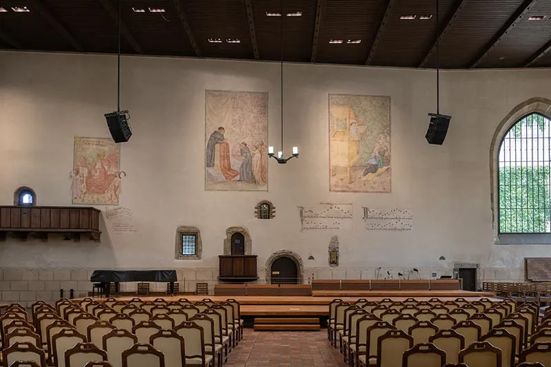 Interior de la Capilla de Belén con hileras de sillas, murales en las paredes y una gran ventana arqueada