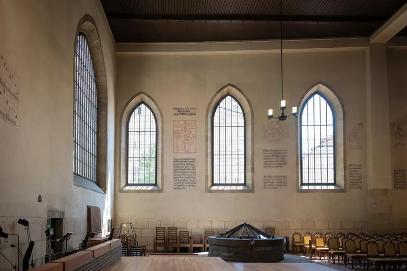 Interior de una capilla con altos ventanales arqueados, muros de piedra y sillas de madera