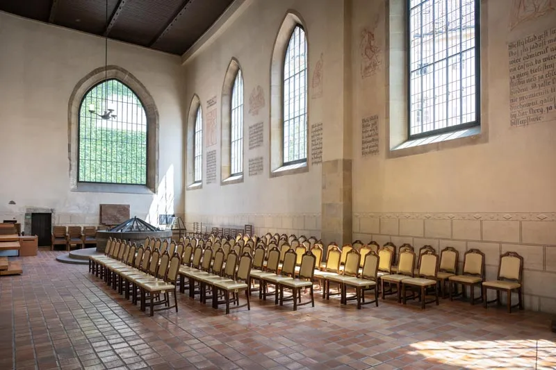 Interior de la Capilla de Belén con filas de sillas, ventanas arqueadas y muros de piedra