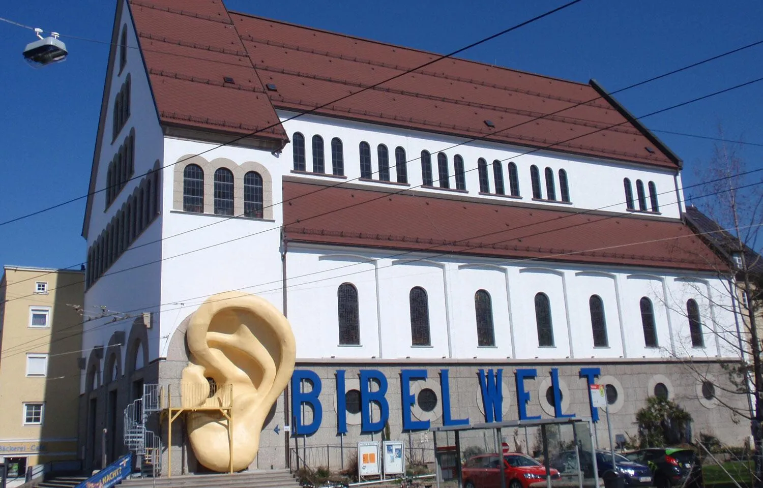 White church building with red-brown roof featuring a large golden ear sculpture and blue BIBELWELT sign in front