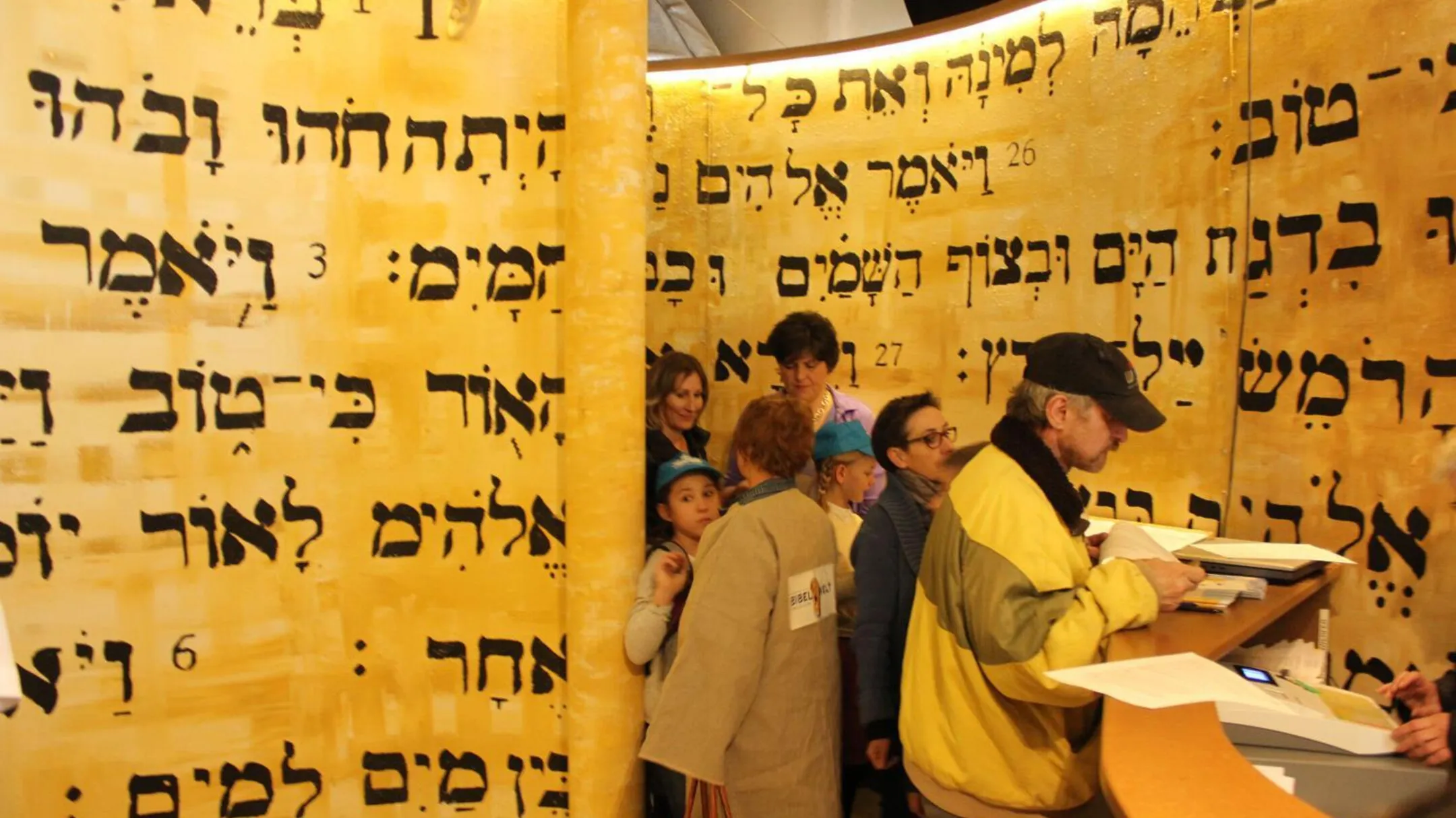 Visitors inside an exhibition space with large golden parchment-like walls covered in black Hebrew text, with a man reading from a book at a wooden lectern in the foreground.