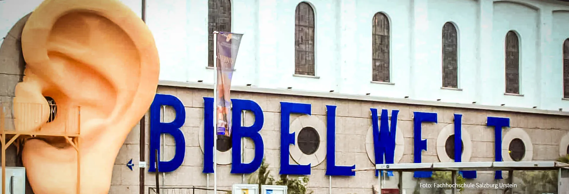 Giant sculptural ear installation beside the blue BIBELWELT lettering on the facade of a church building with arched windows