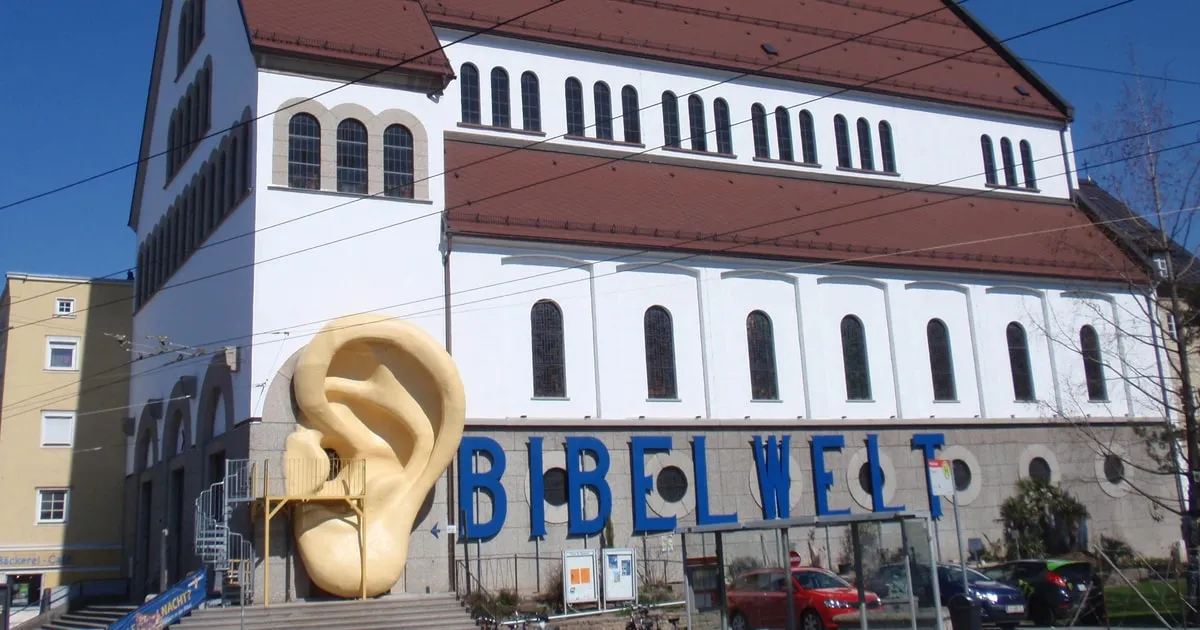 White church building facade with a large beige ear sculpture in front and blue BIBELWELT lettering on the wall