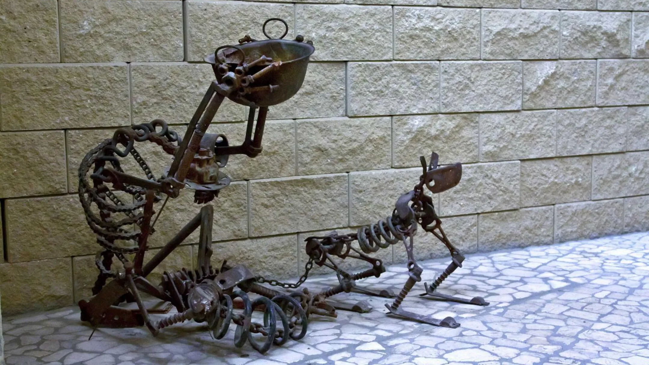 Metal sculptures of a human skeleton and a dog skeleton made from scrap metal and tools, displayed against a stone wall.