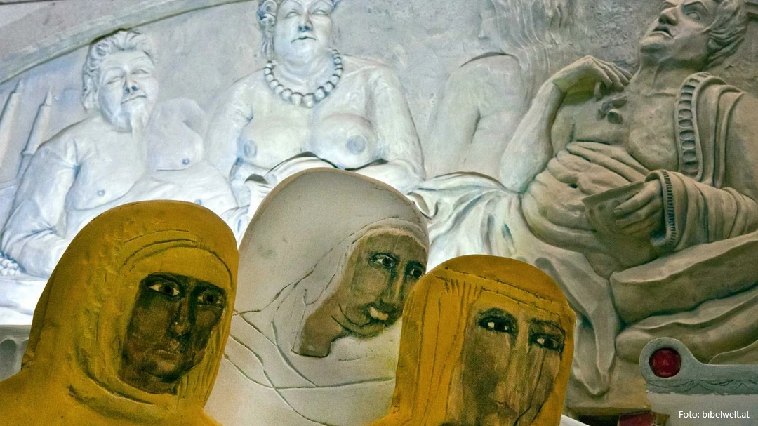 Sculptural exhibition installation with three stylized yellow and white foreground heads in front of a white classical relief carving showing multiple figures including a central female figure with a necklace
